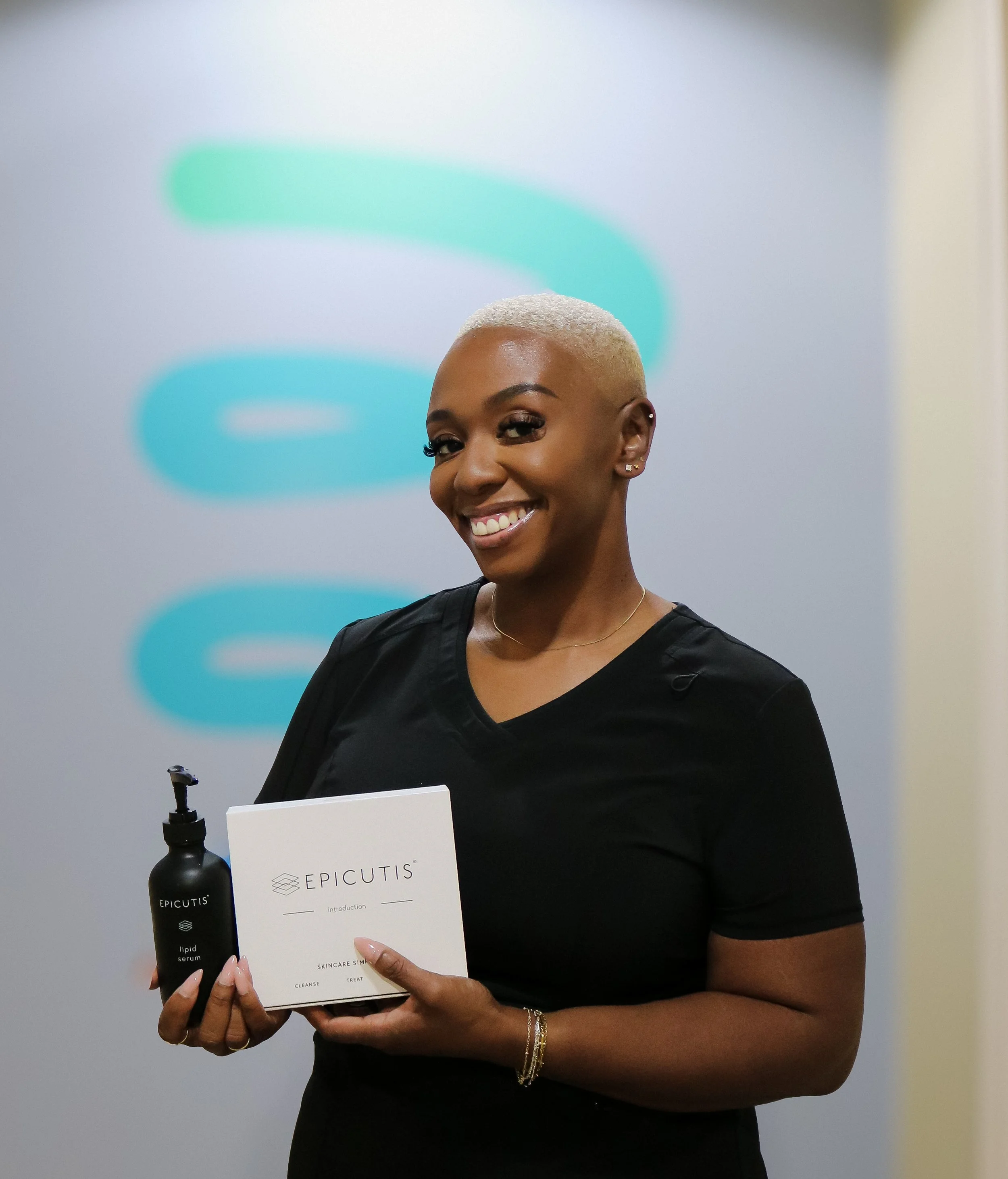 Woman with short blonde hair, wearing a black shirt and jewelry, smiling and holding a box labeled 'EPICUTIS' and a black bottle of serum, standing in front of a gray wall with colorful abstract shapes.
