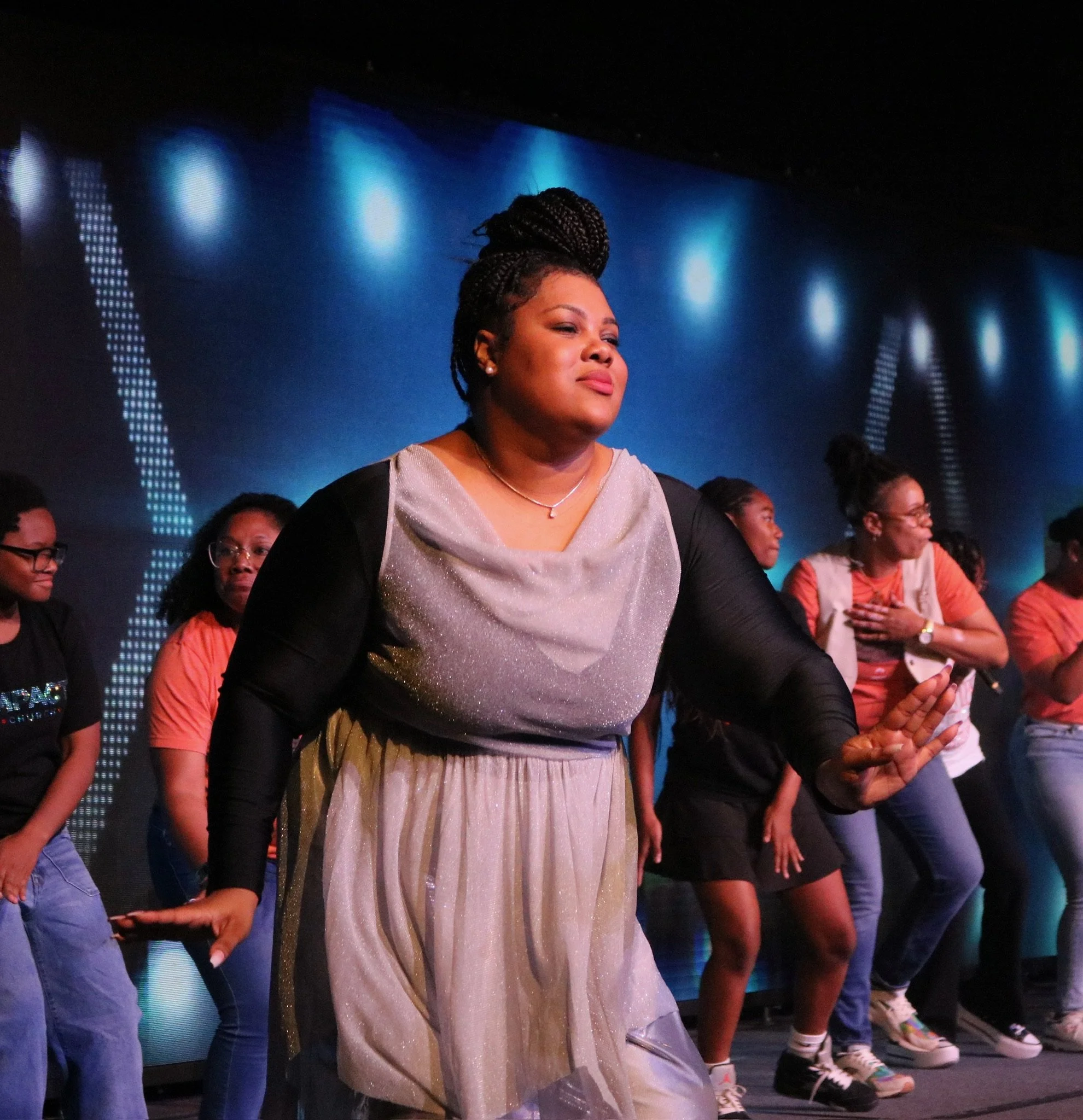 Woman with braided hair standing among a group of diverse people on stage, with blue lighting and digital patterns on a screen in the background.