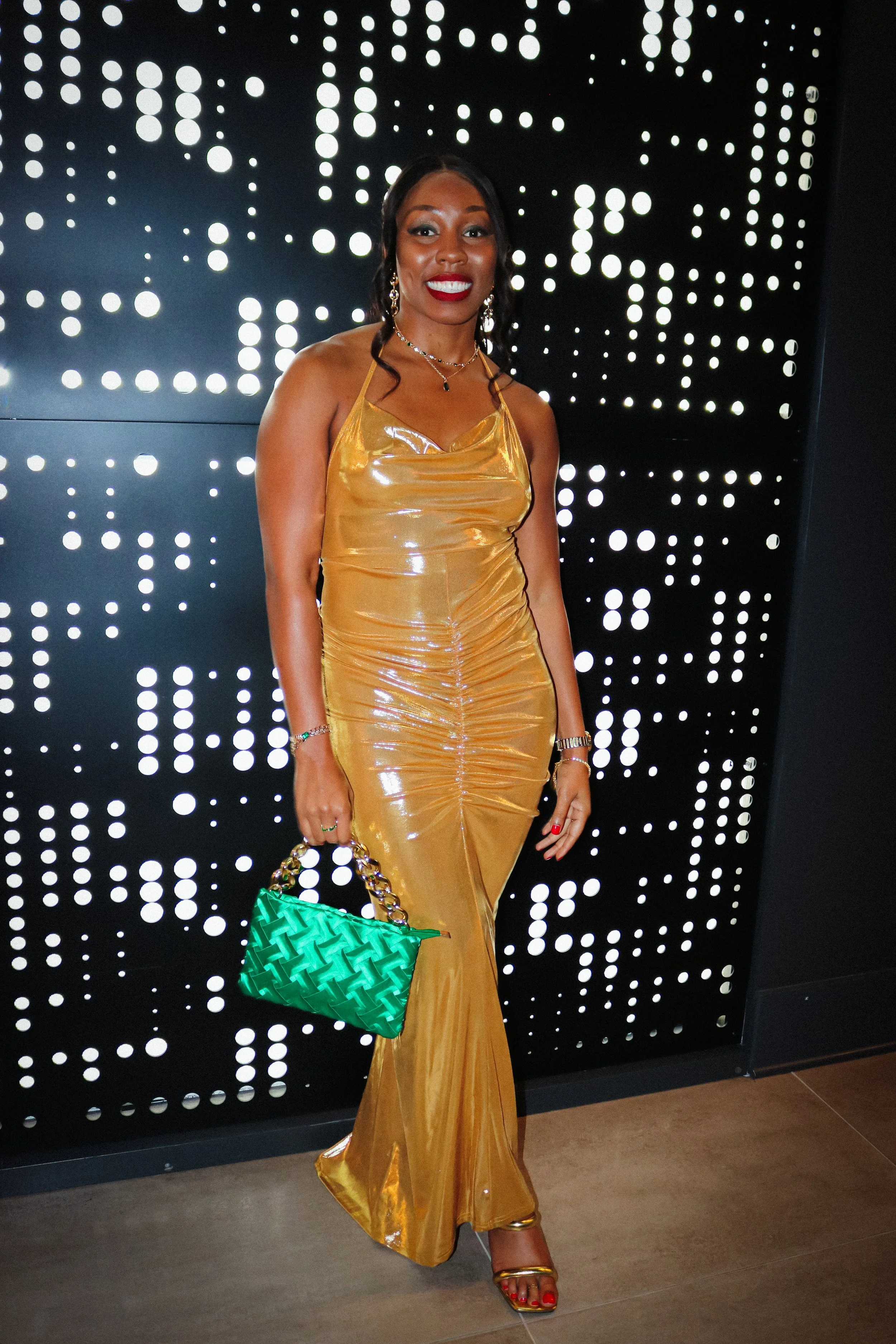 A woman wearing a shiny, gold evening gown, standing in front of a black wall with decorative white circular light patterns. She is holding a green quilted handbag with a gold chain strap, and wearing jewelry including earrings, necklaces, and bracelets, with short, styled black hair and red lipstick.