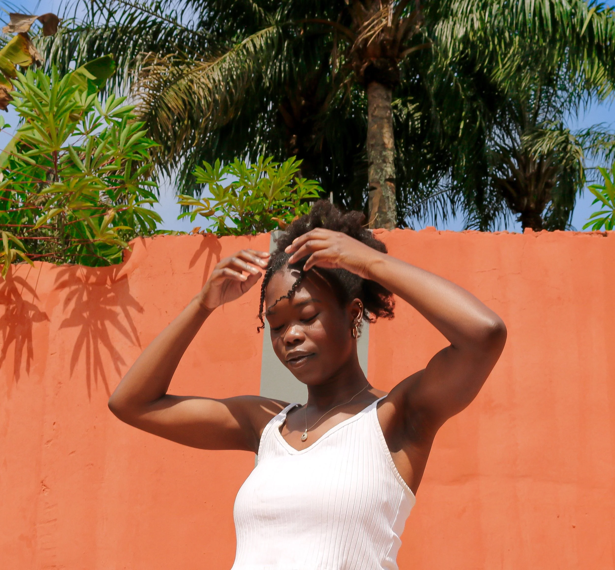 A woman with curly hair and earrings standing outdoors in front of an orange wall, adjusting her hair with both hands, wearing a white sleeveless top, with lush green palm and leafy plants, and a blue sky in the background.
