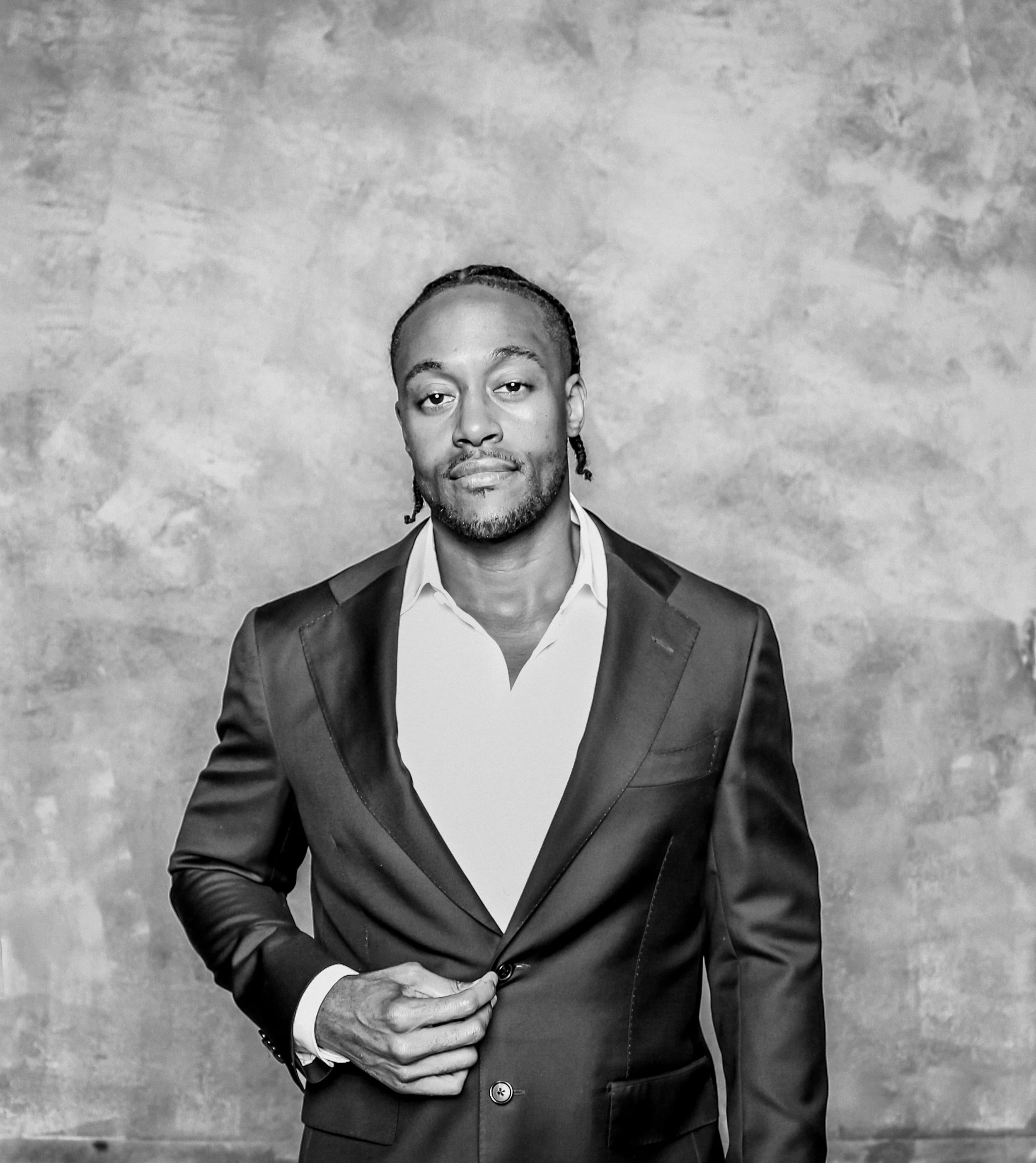 Black and white photograph of a man in a suit, standing against a textured background. He is looking at the camera with a neutral expression, with his right hand holding the sleeve of his left hand at waist level.