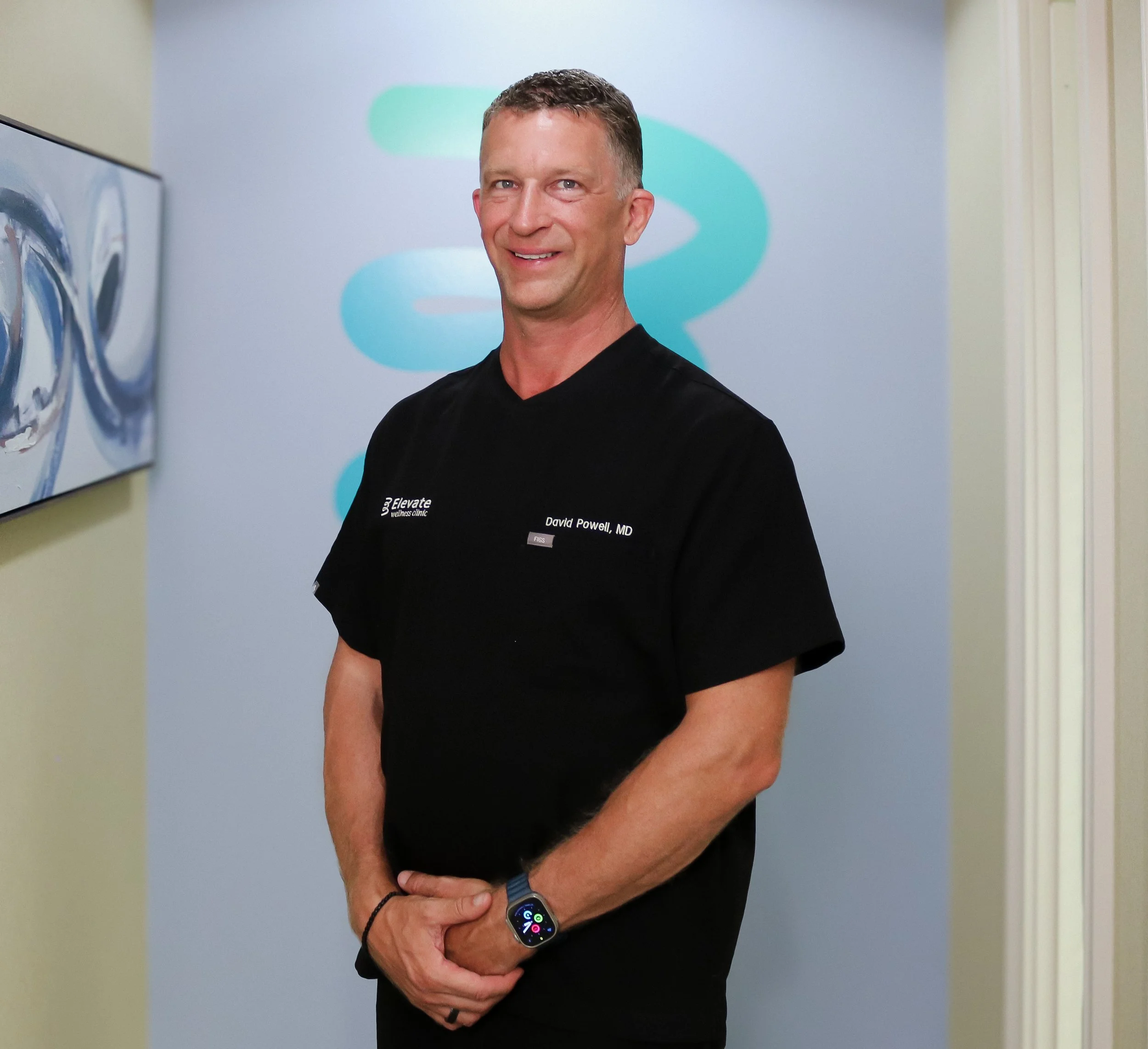 A man with short hair and a smile, wearing a black shirt with medical branding, standing against a light-colored wall with abstract art in the background, and wearing a smartwatch.