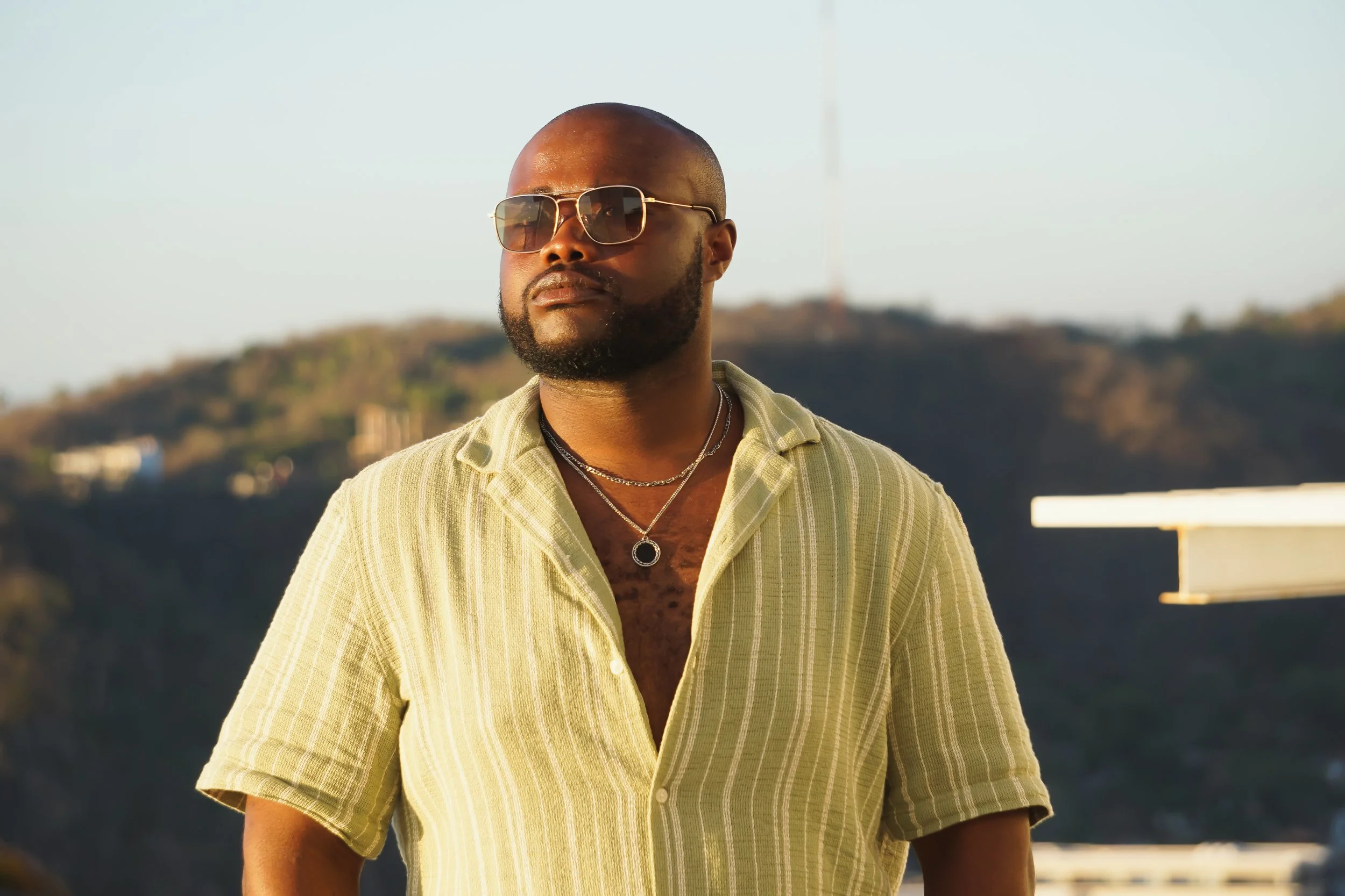 A man with a beard, wearing aviator sunglasses, a yellow plaid shirt, and layered silver necklaces, standing outdoors with a blurry natural landscape in the background during sunlight.