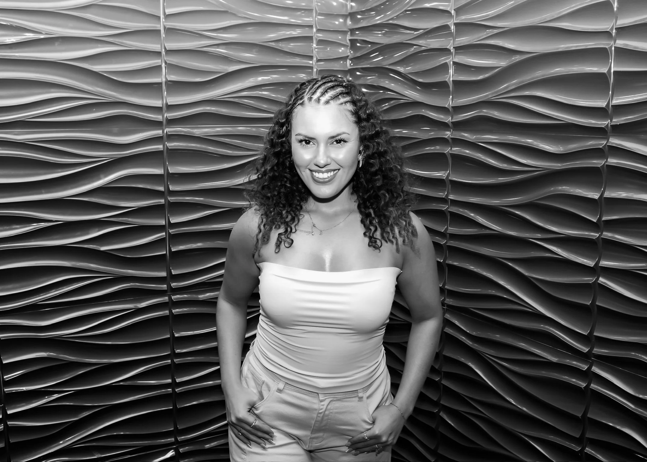 A smiling woman with curly hair wearing a strapless top and light-colored pants standing in front of an abstract, textured wall with curved patterns.