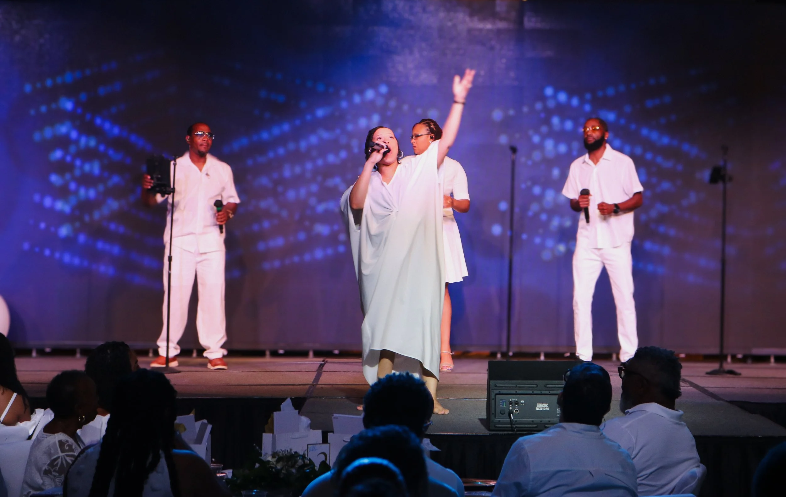 A woman dressed in white, holding a microphone and singing on stage with her arm raised, accompanied by three men also in white, holding microphones and standing in front of a digital blue dotted background.