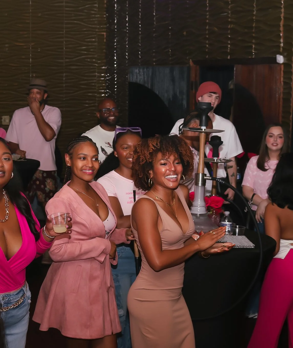 Group of smiling people at a social gathering, some holding drinks, with a hookah on a table in the foreground.