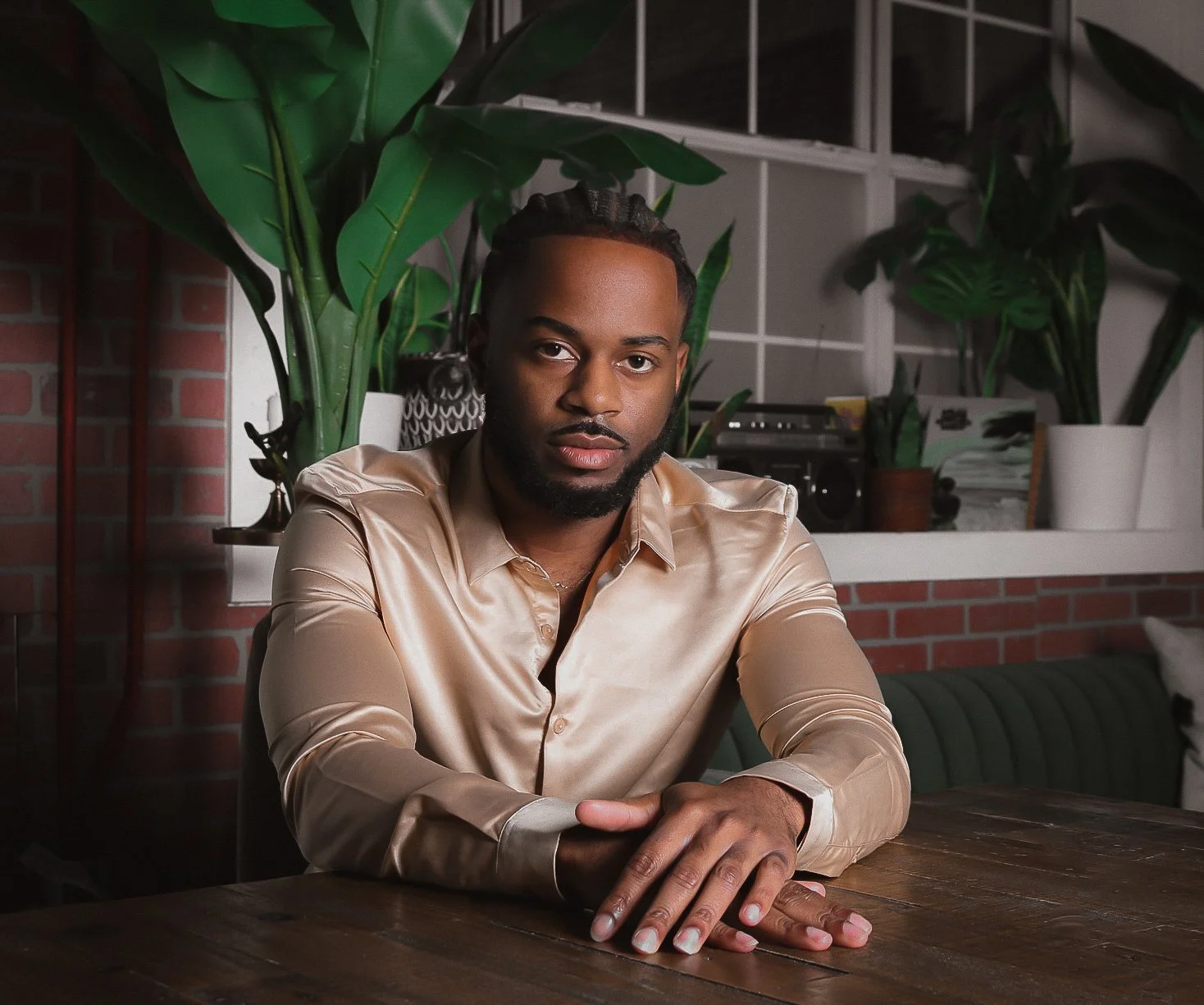 A man with braids and a beard, wearing a shiny beige silk shirt, sitting at a wooden table in a room with indoor plants and a brick wall background.