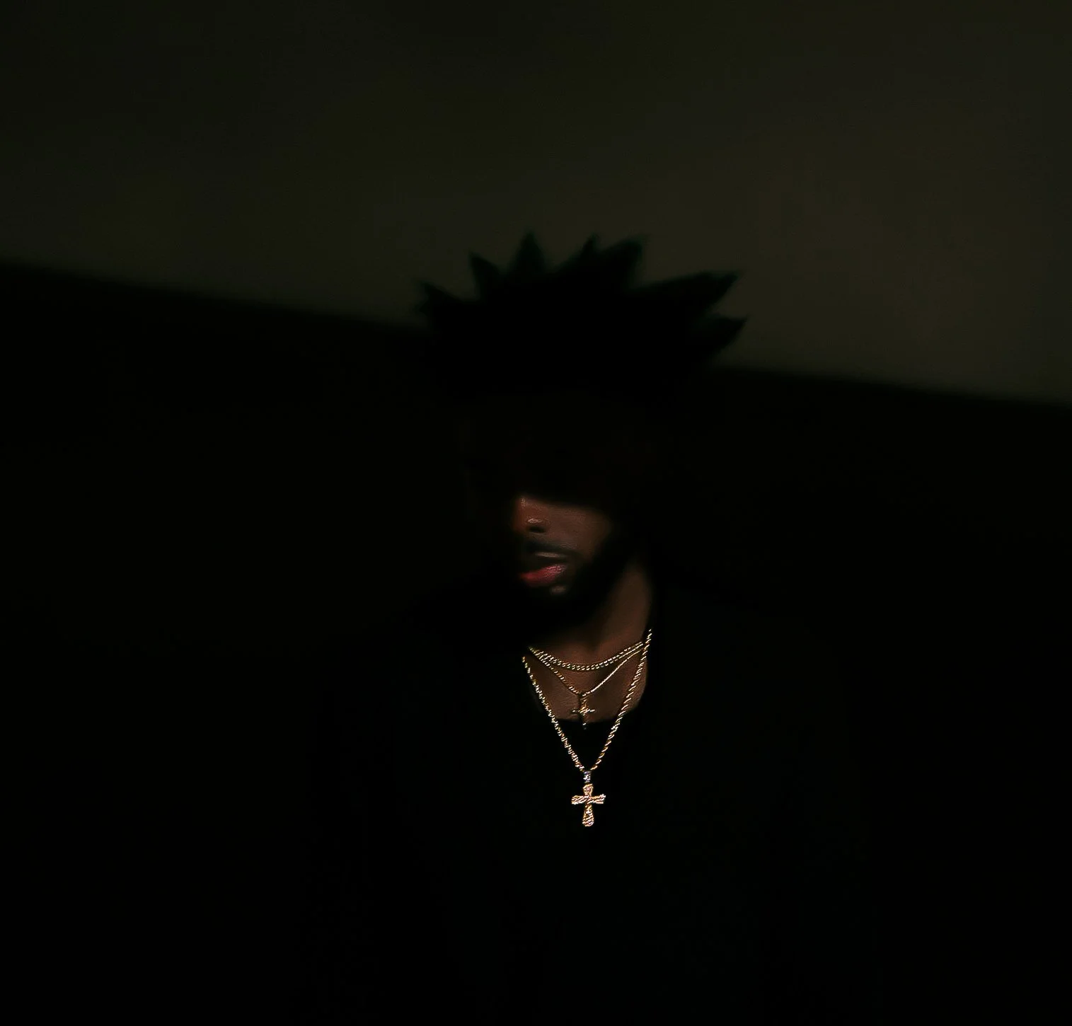Low-light photo of a person with a large spiked hairstyle, wearing multiple layered necklaces, including a cross pendant, and a dark top against a dark background.