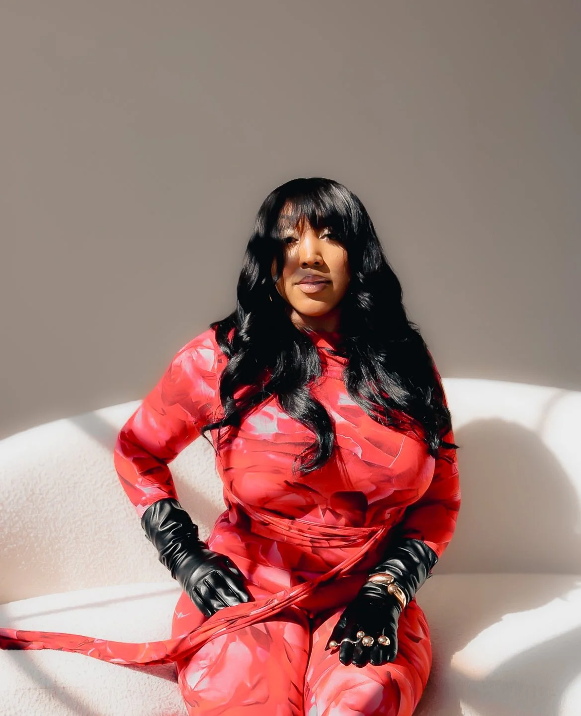 Woman with long black hair in loose waves, dressed in a red patterned dress and black gloves, sitting on a white couch in natural light.
