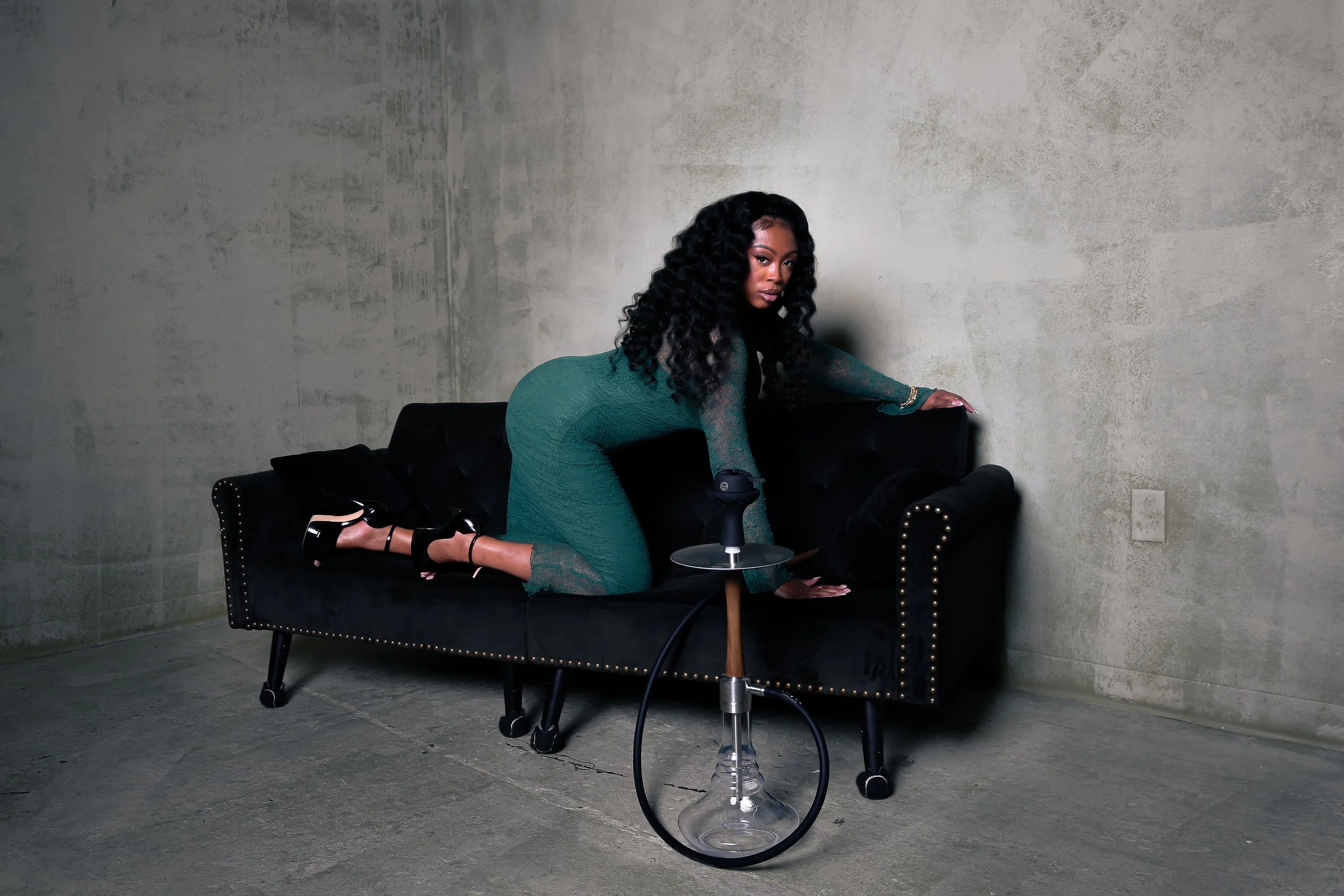 A woman with long curly black hair wearing a fitted green dress and high heels is positioned on a black velvet sofa with brass nailhead trim. She is leaning forward with her hands on the sofa and knees on the seat, looking towards the camera. The setting is a minimalistic room with a light gray, slightly textured wall and concrete floor, featuring a small modern glass table with a hookah on it in front of the sofa.