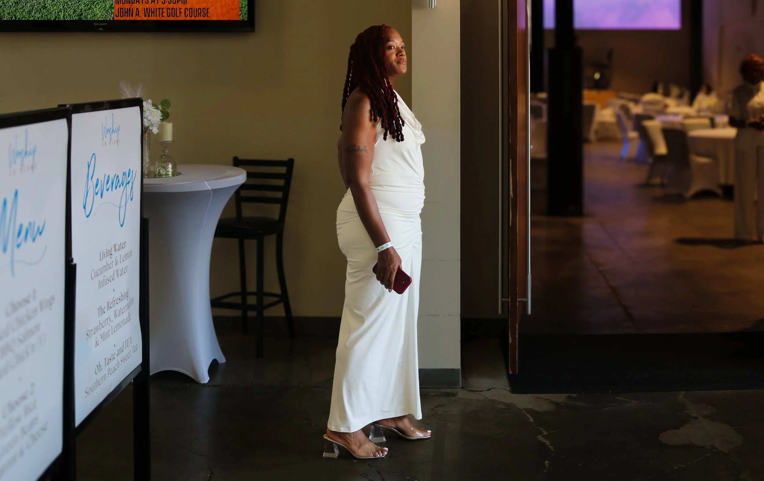 A woman with dreadlocks wearing a white dress and transparent heels standing near an entrance, holding a red phone, inside a banquet hall or event space with tables and chairs in the background.