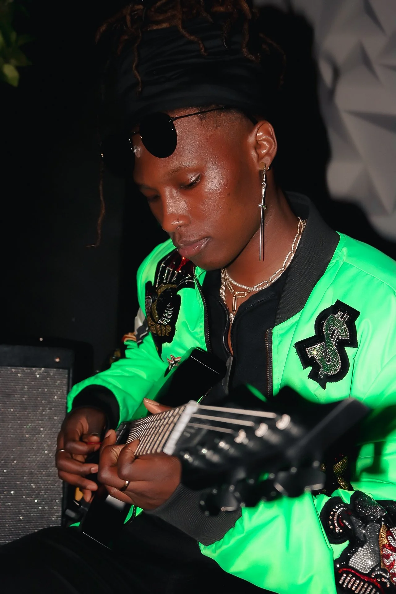 A person with dark skin, sporting dreadlocks with white tips, wearing a black cap and large round sunglasses, dressed in a bright green jacket with embellishments, holding a musical instrument or equipment, sitting indoors.