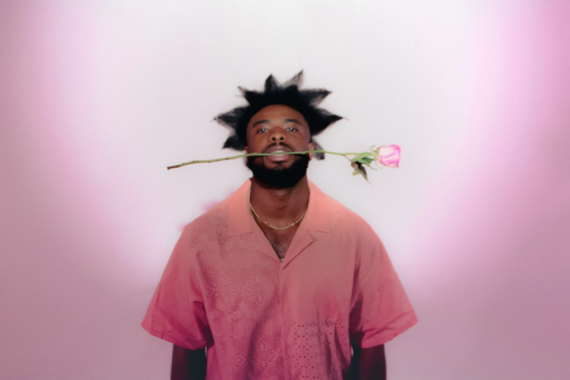 A man with dark skin, facial hair, and spiky hair holding a pink rose in his mouth, wearing a pink shirt and a gold necklace, standing against a pink background.
