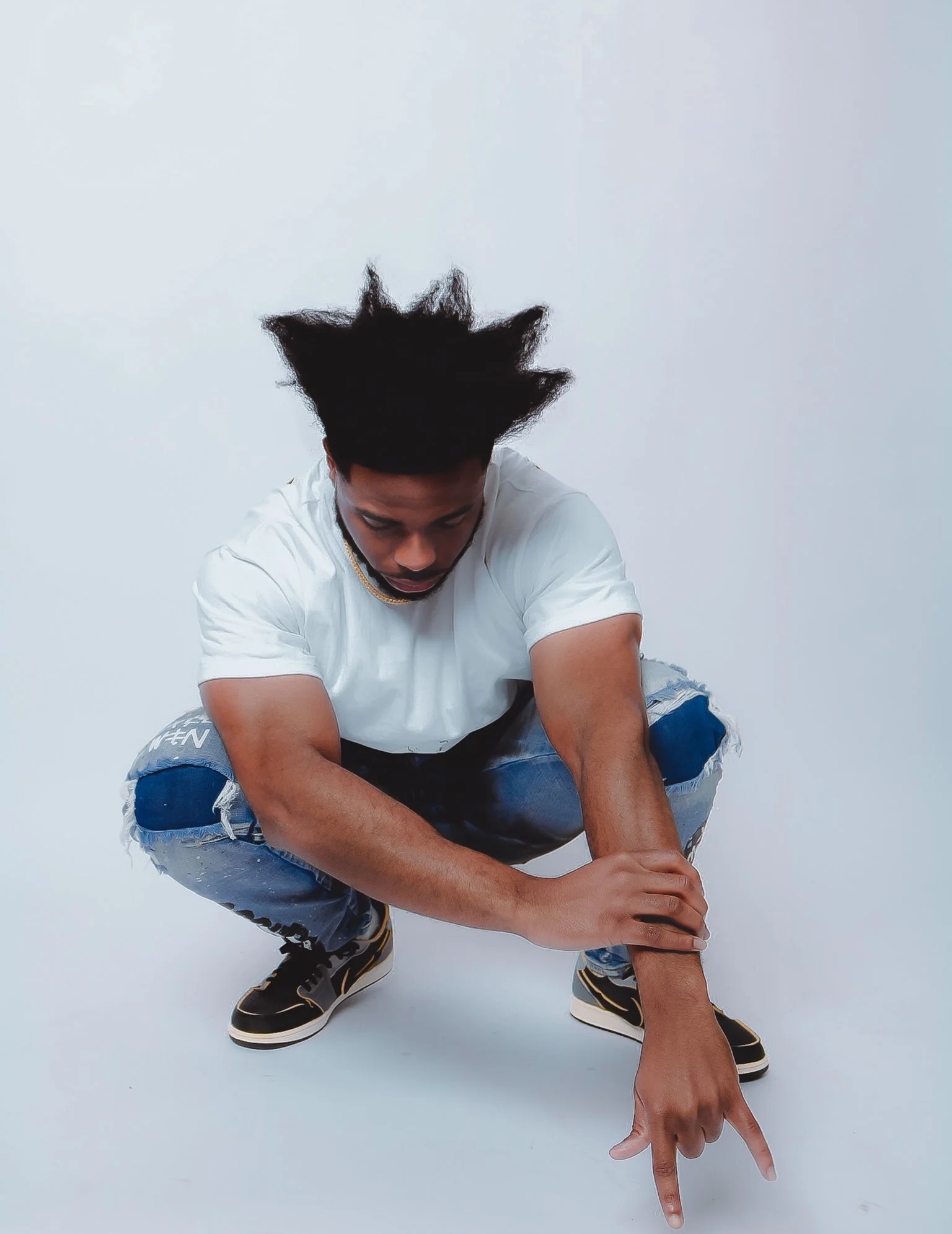 A young man with styled short black hair, wearing a white t-shirt, ripped jeans, and black sneakers with white soles, crouching down with one hand on his knee and the other hand pointing with two fingers, set against a light gray background.