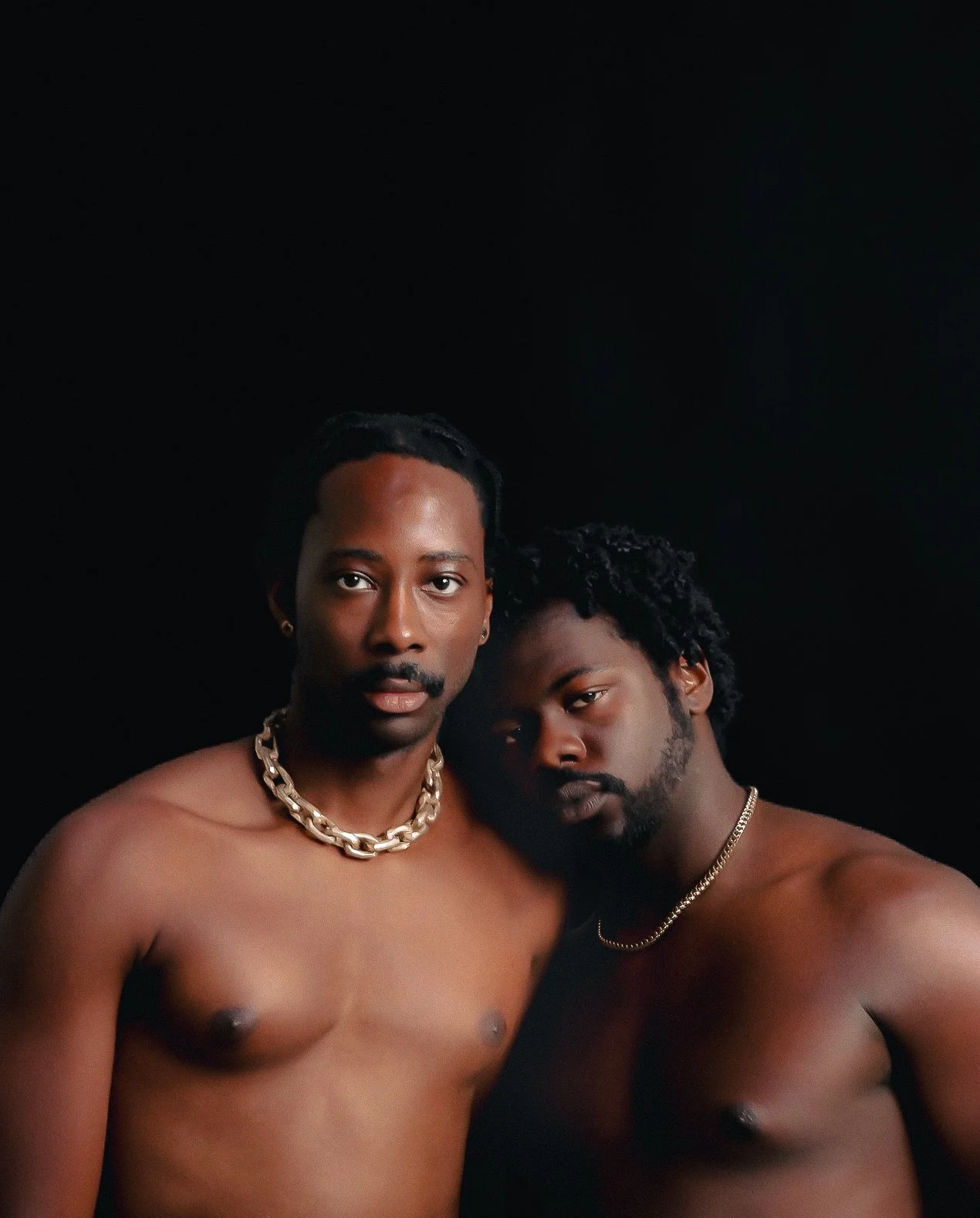 Two shirtless Black men with necklaces posing against a black background. One has shockingly close-cropped hair, a gold chain, and a mustache. The other has dreadlocks, a gold chain, and a beard, with his head leaning on the first man's shoulder.