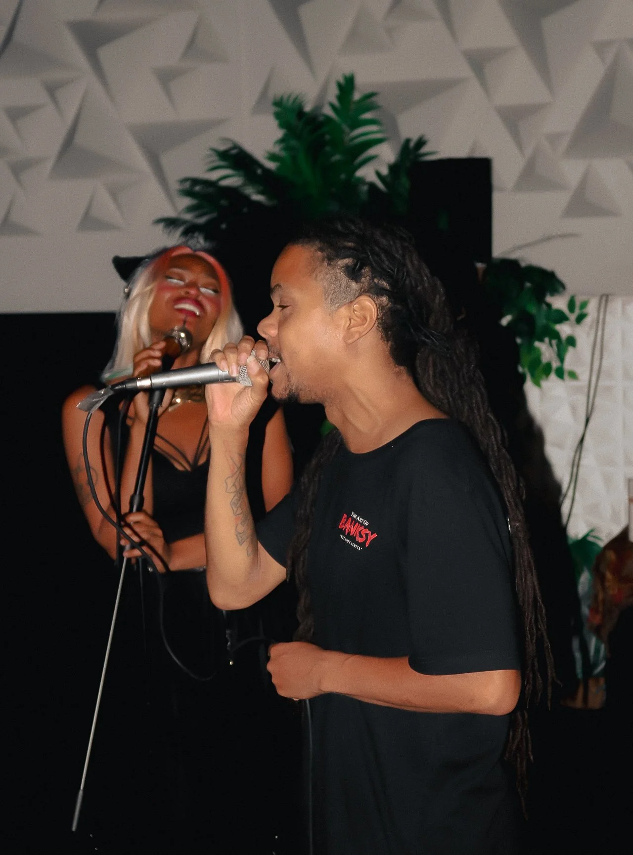 A man with dreadlocks singing into a microphone while a woman with platinum blonde hair and makeup listens nearby in a room with a patterned ceiling and plants.