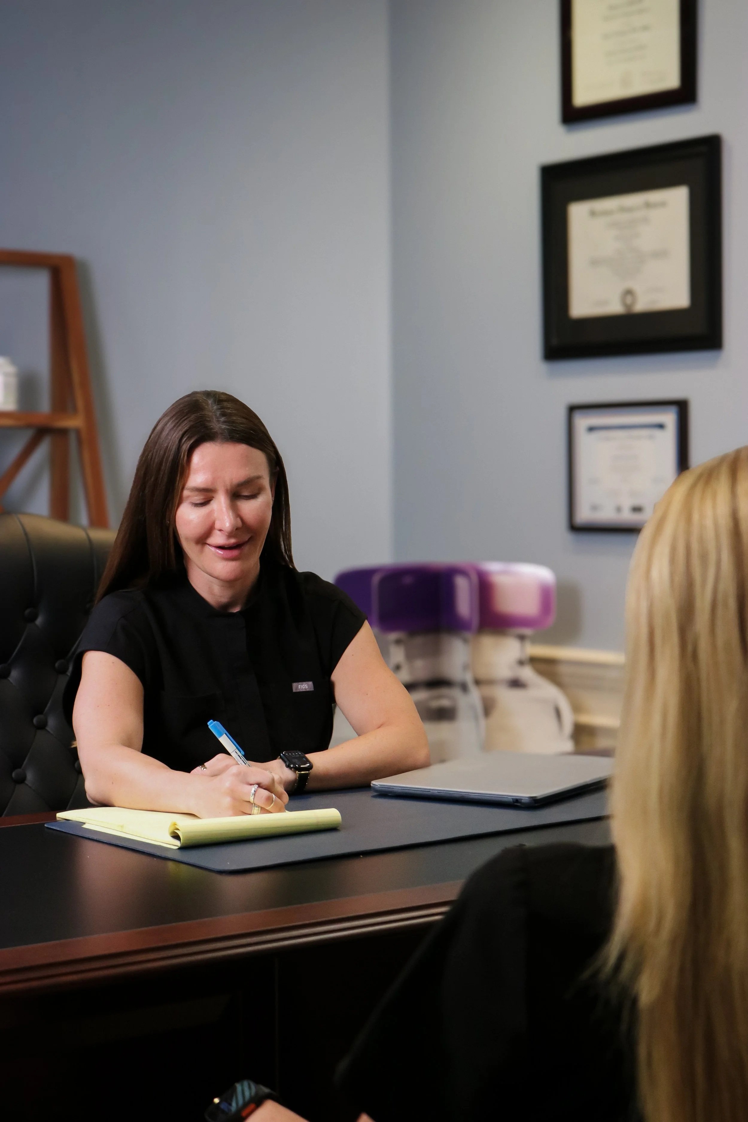 A woman with long brown hair wearing a black shirt is sitting at a desk, smiling, and writing in a yellow notebook. There are framed certificates on the wall behind her, and a purple high chair in the background.