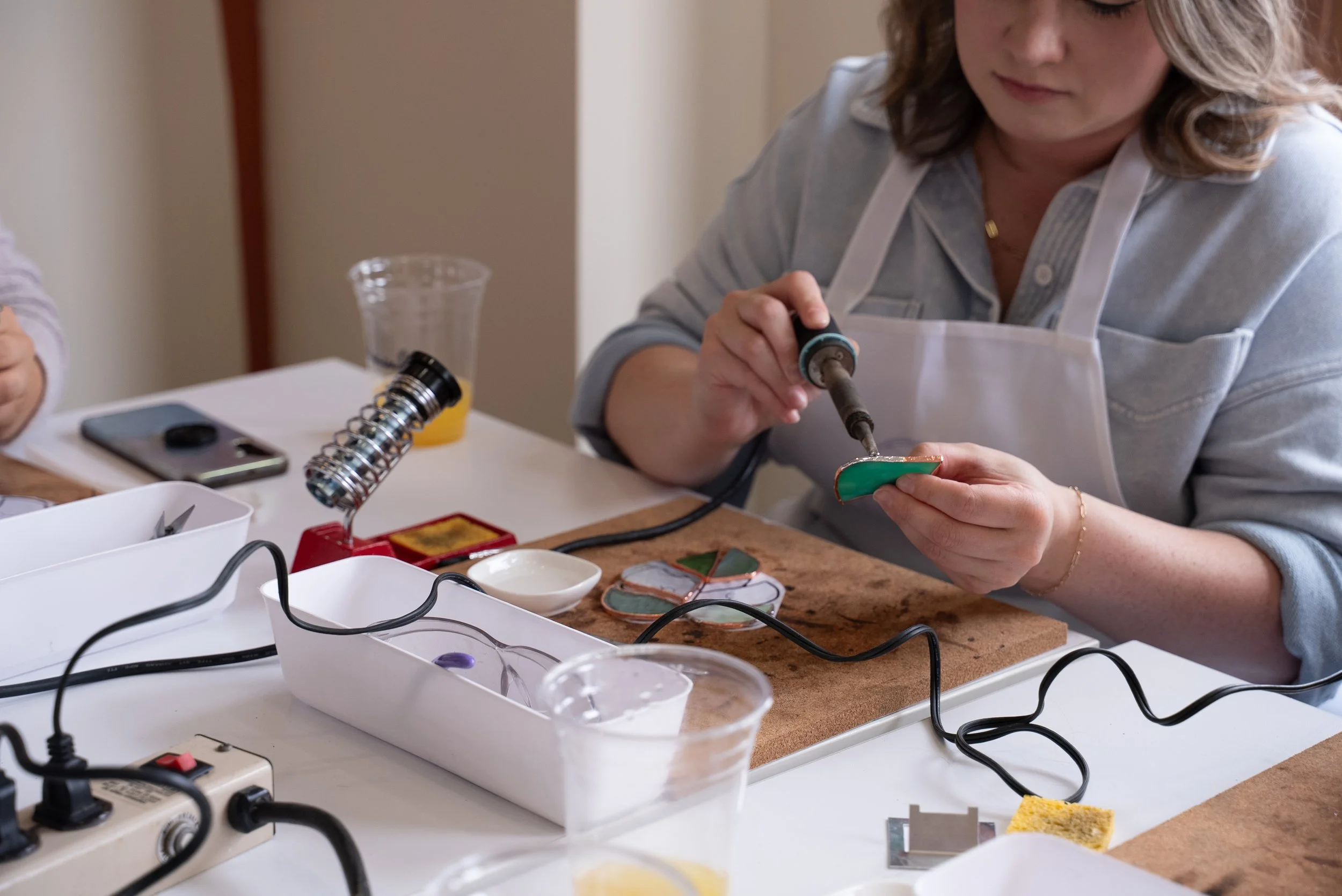 Woman soldering a small green object at a workbench with various tools and supplies around her.