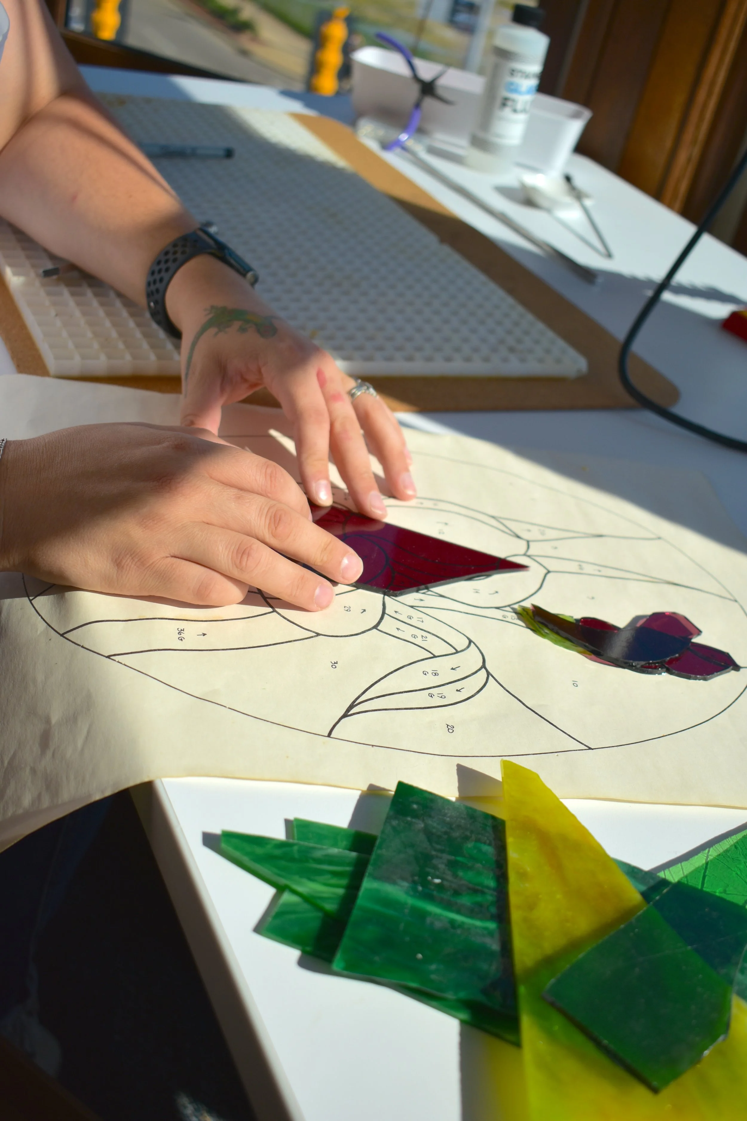 A person working on a paint-by-number craft, with color samples in green and yellow in the foreground, and various supplies and tools on the table. The person is wearing a black watch and has a tattoo on their wrist.