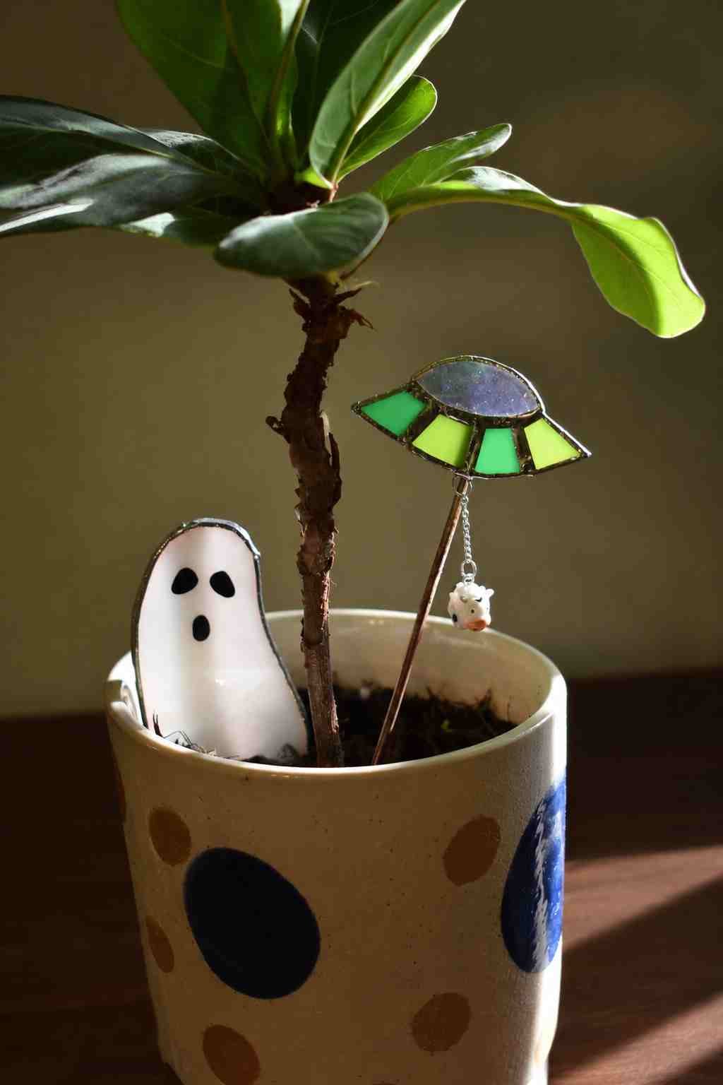 A potted plant with green leaves decorated with Halloween-themed ornaments, including a ghost figurine, a stained glass umbrella, and a small bear charm.