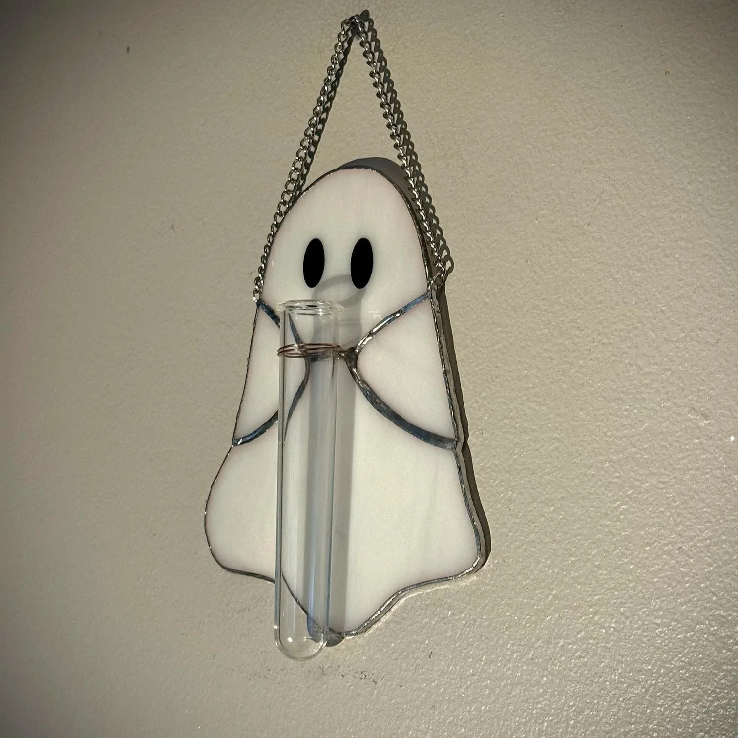 A stained glass ghost holding a propagation tube with wire. The ghost is hung on a wall with a chain.