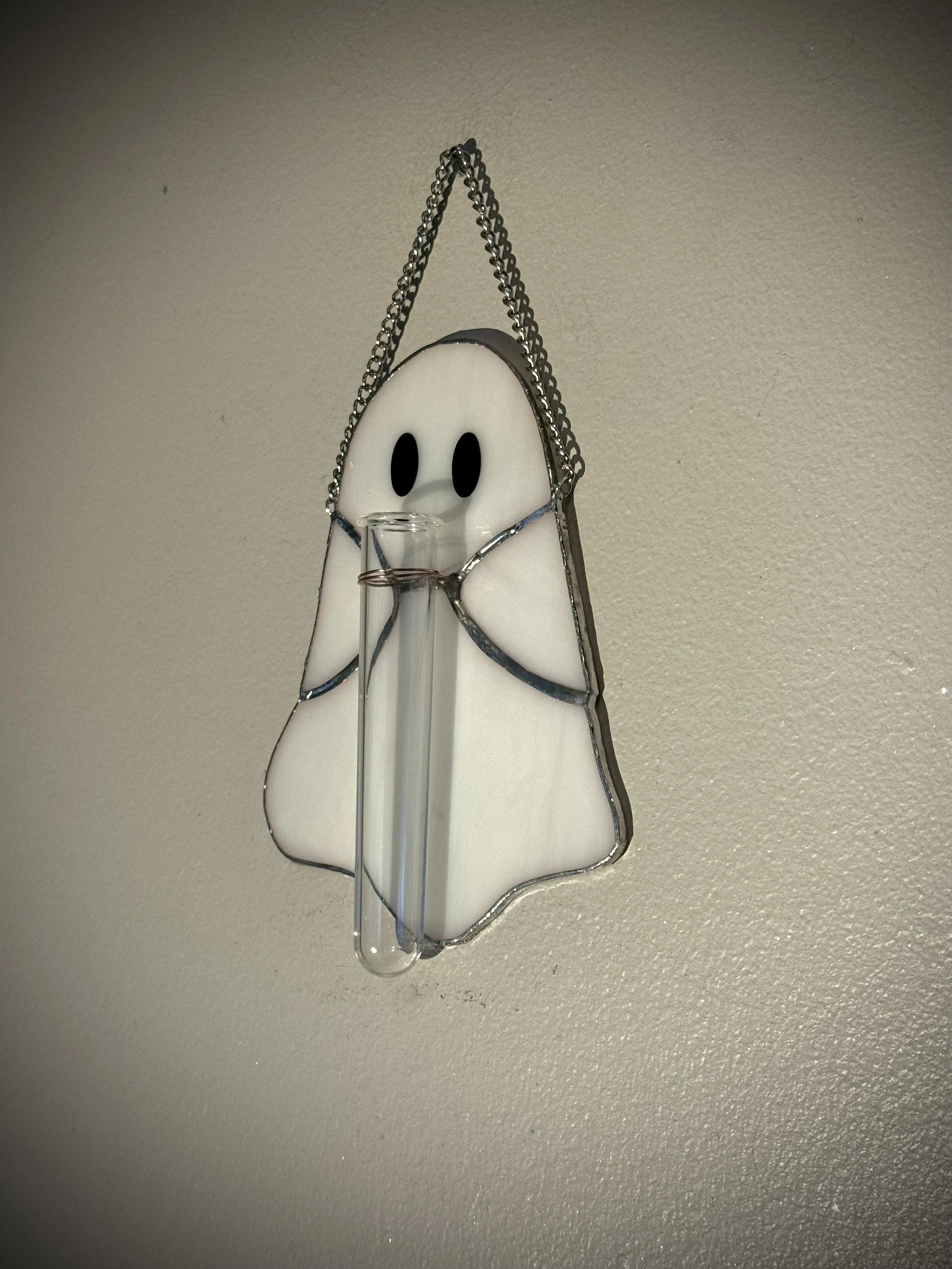 A decorative wall hanging shaped like a ghost with a chain hanger, capturing a light source that creates a stained glass effect, showing a ghost's face with black eyes and a small nose.