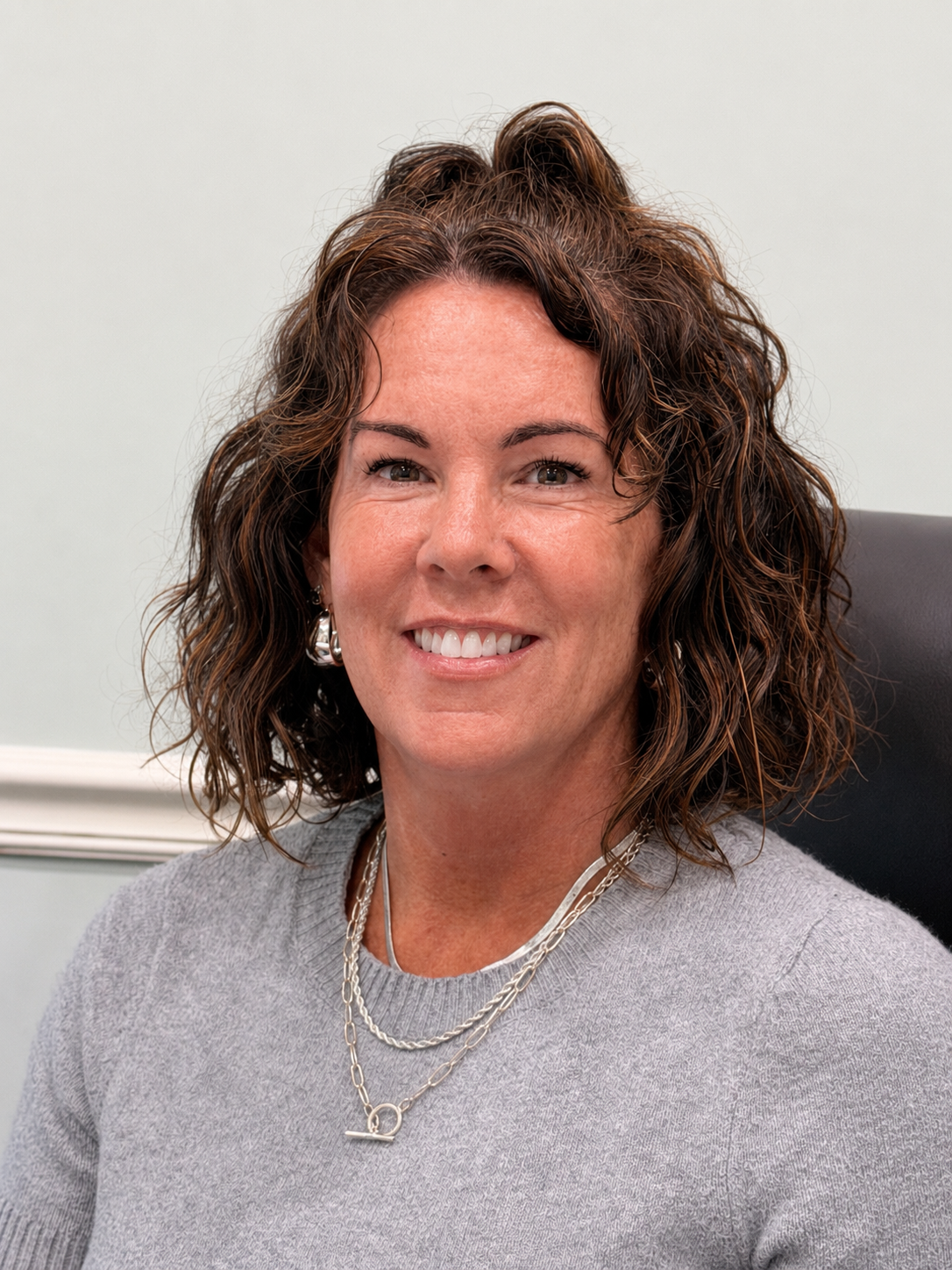 A woman with shoulder-length curly brown hair smiling, sitting in a black chair, wearing a light gray sweater and layered silver necklaces.
