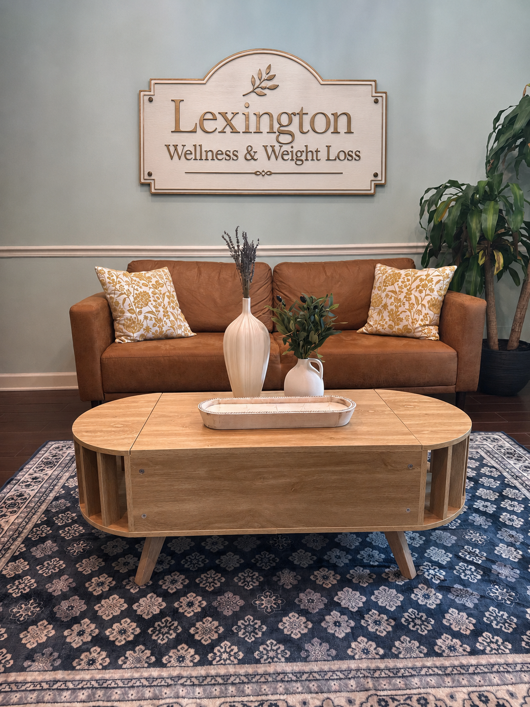 Reception area of Lexington Wellness & Weight Loss with a brown leather couch, decorative pillows, wooden coffee table with vases and greenery, potted plant, and a sign on the wall reading 'Lexington Wellness & Weight Loss'.