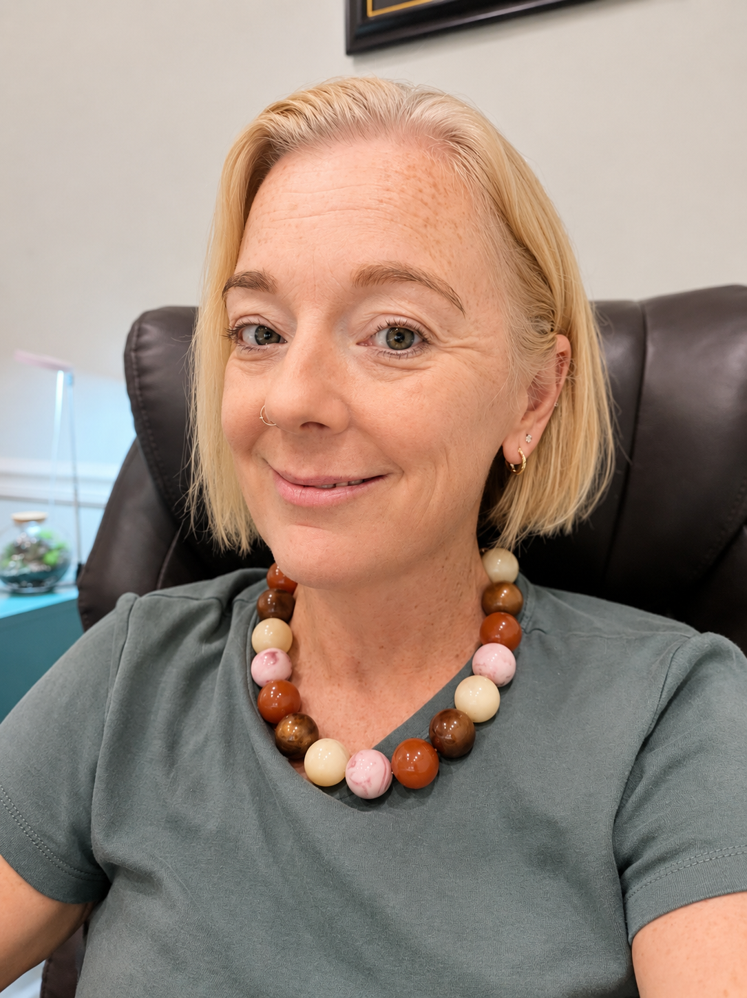 A woman with blonde hair sitting in a black leather chair, smiling at the camera, wearing a gray shirt, large beaded necklace in shades of brown, pink, and cream, with multiple earrings, in an office setting.