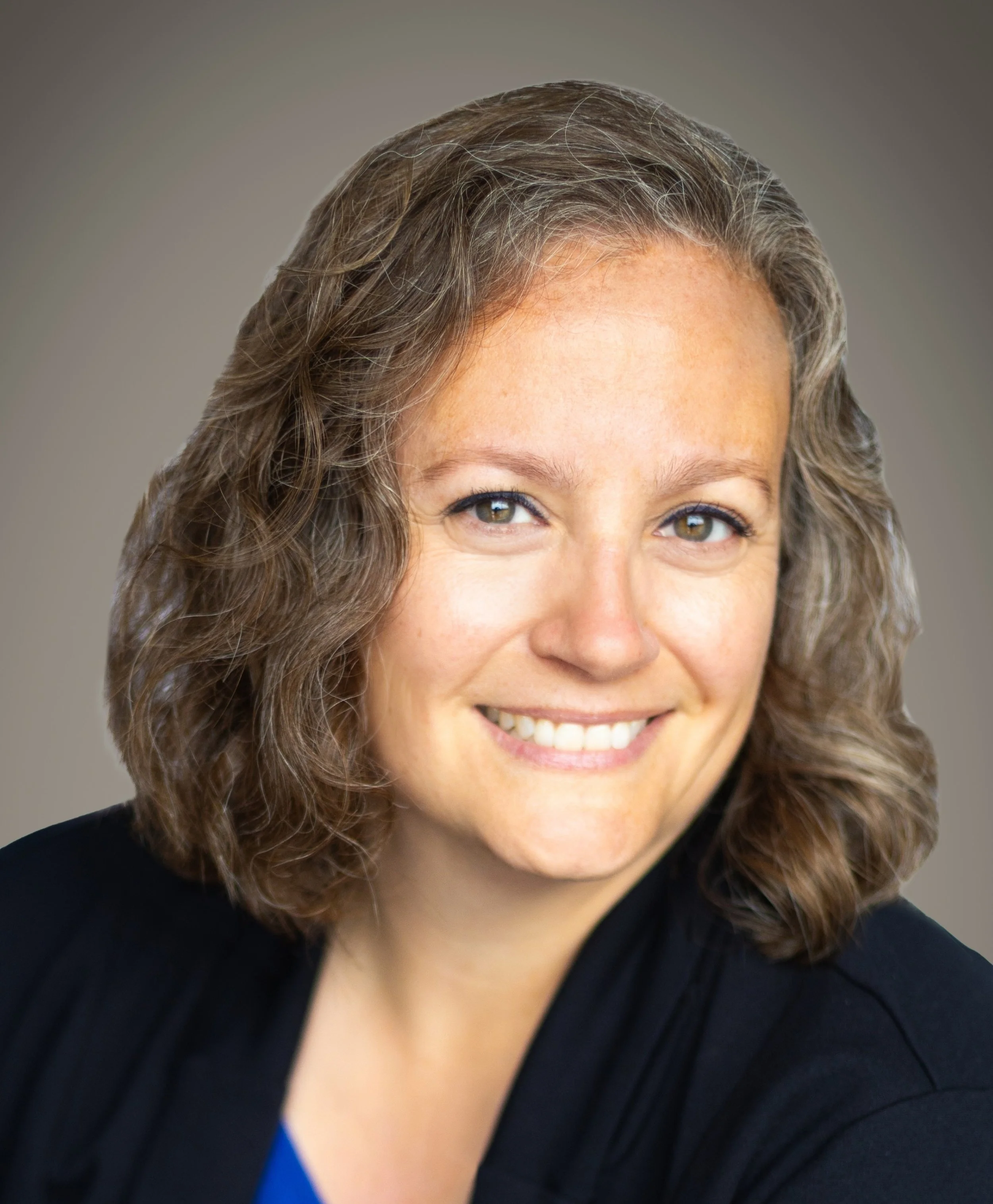 Close-up of a woman with curly brown hair and hazel eyes, smiling, wearing a black blazer with a blue top underneath.