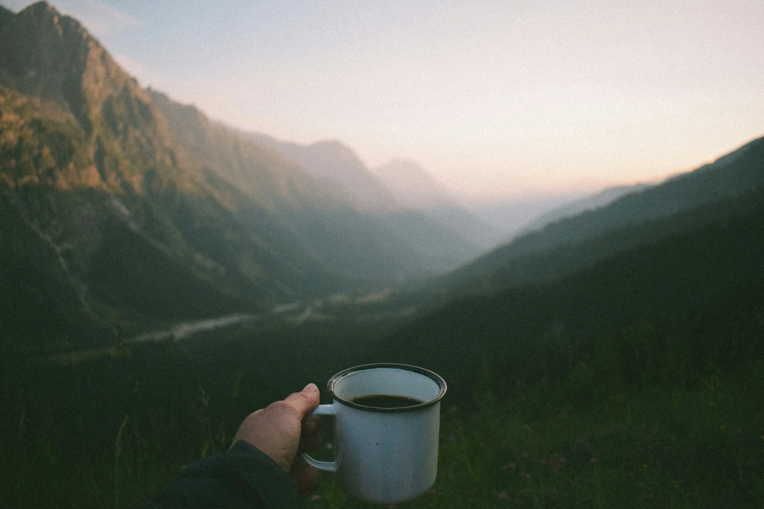 Person holding a white mug of coffee or tea with a mountain landscape in the background, during sunset or sunrise.
