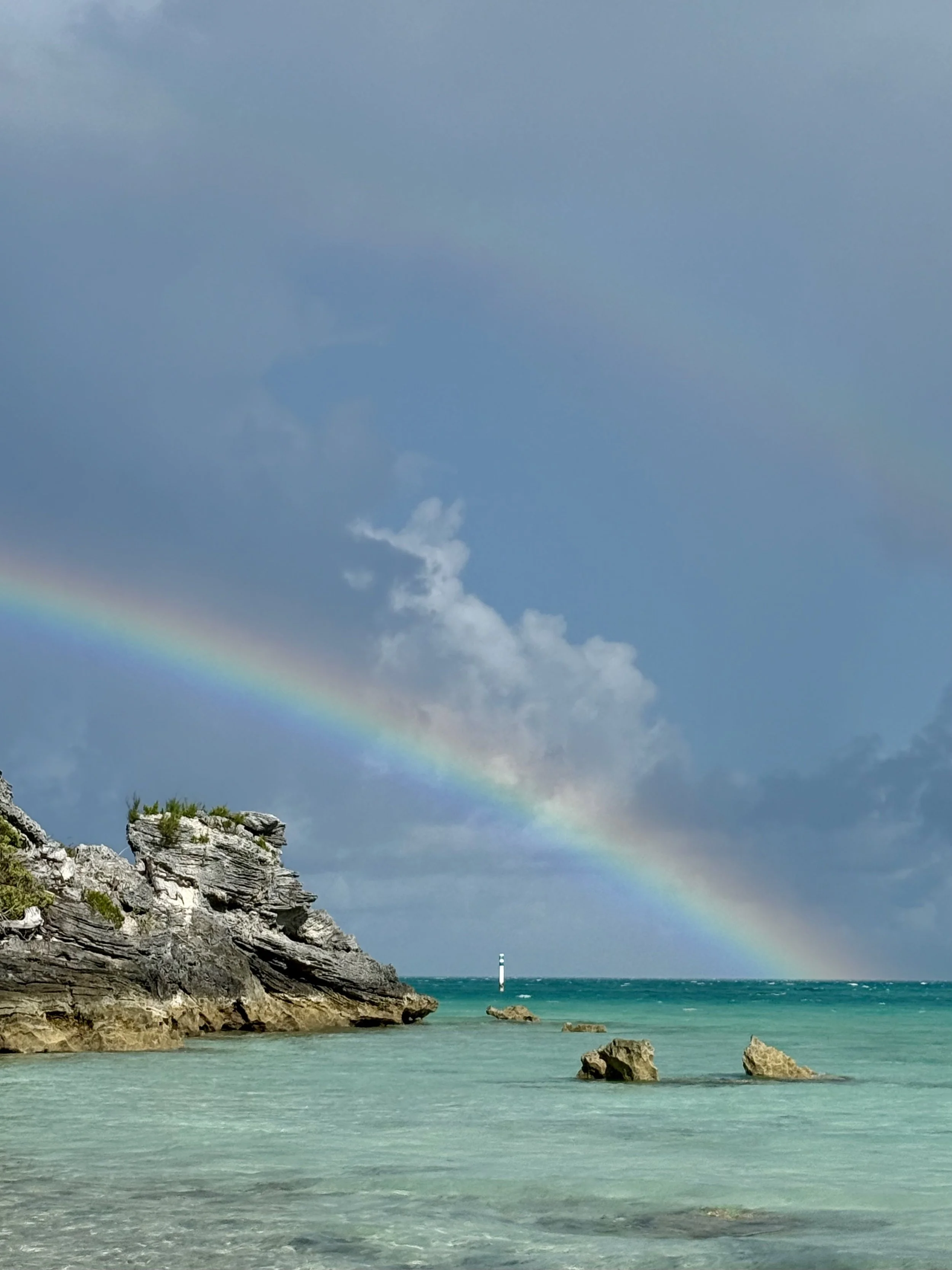 A coastal scene with a rainbow arching across a partly cloudy sky, rocky cliffs on the shore, and a lighthouse in the distance over the ocean.