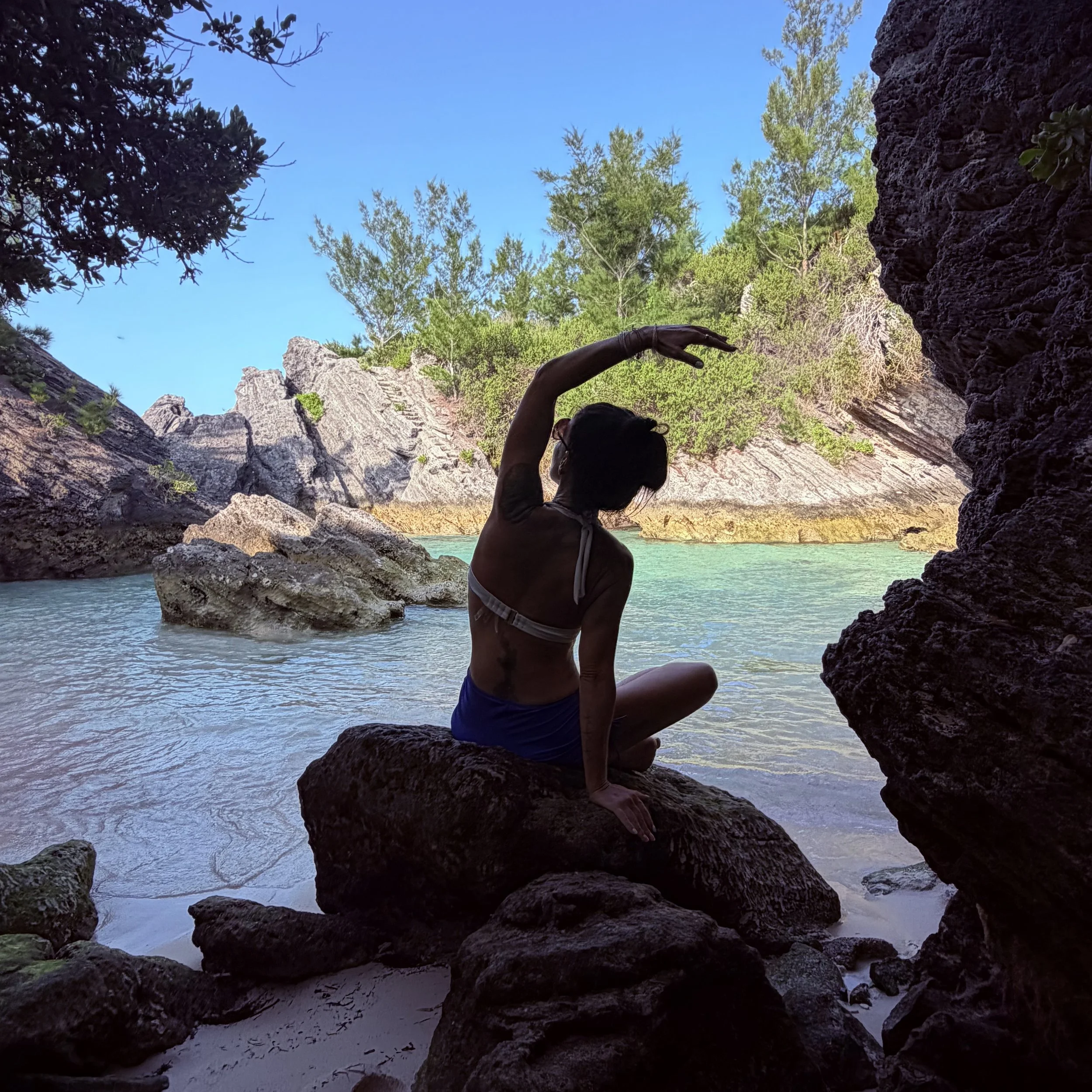 A person sitting on rocks by the water in a canyon with cliffs and trees, reaching their hand towards the sky.