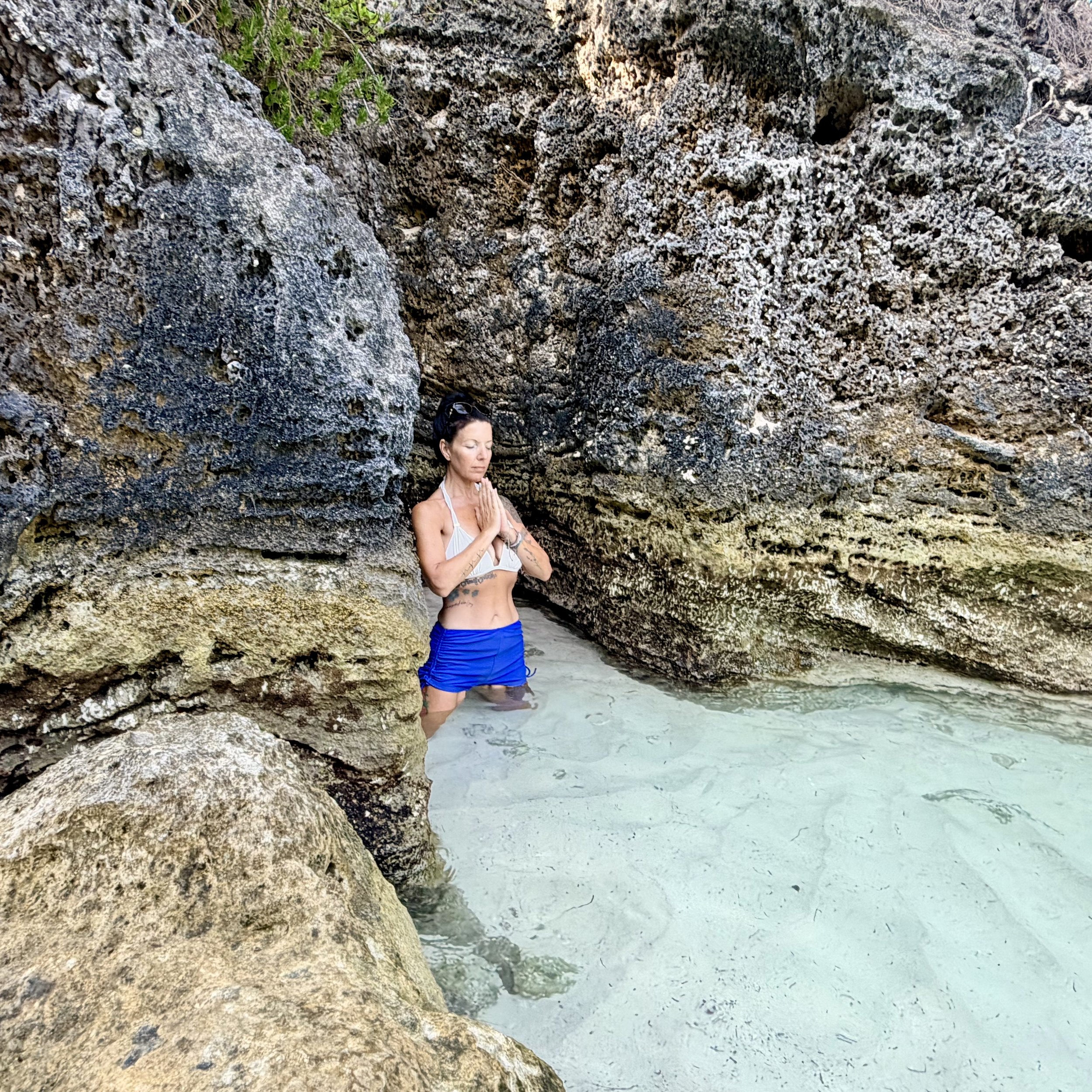 A woman standing in front of large, textured rocks and a body of water, with her eyes closed and hands in a prayer position.