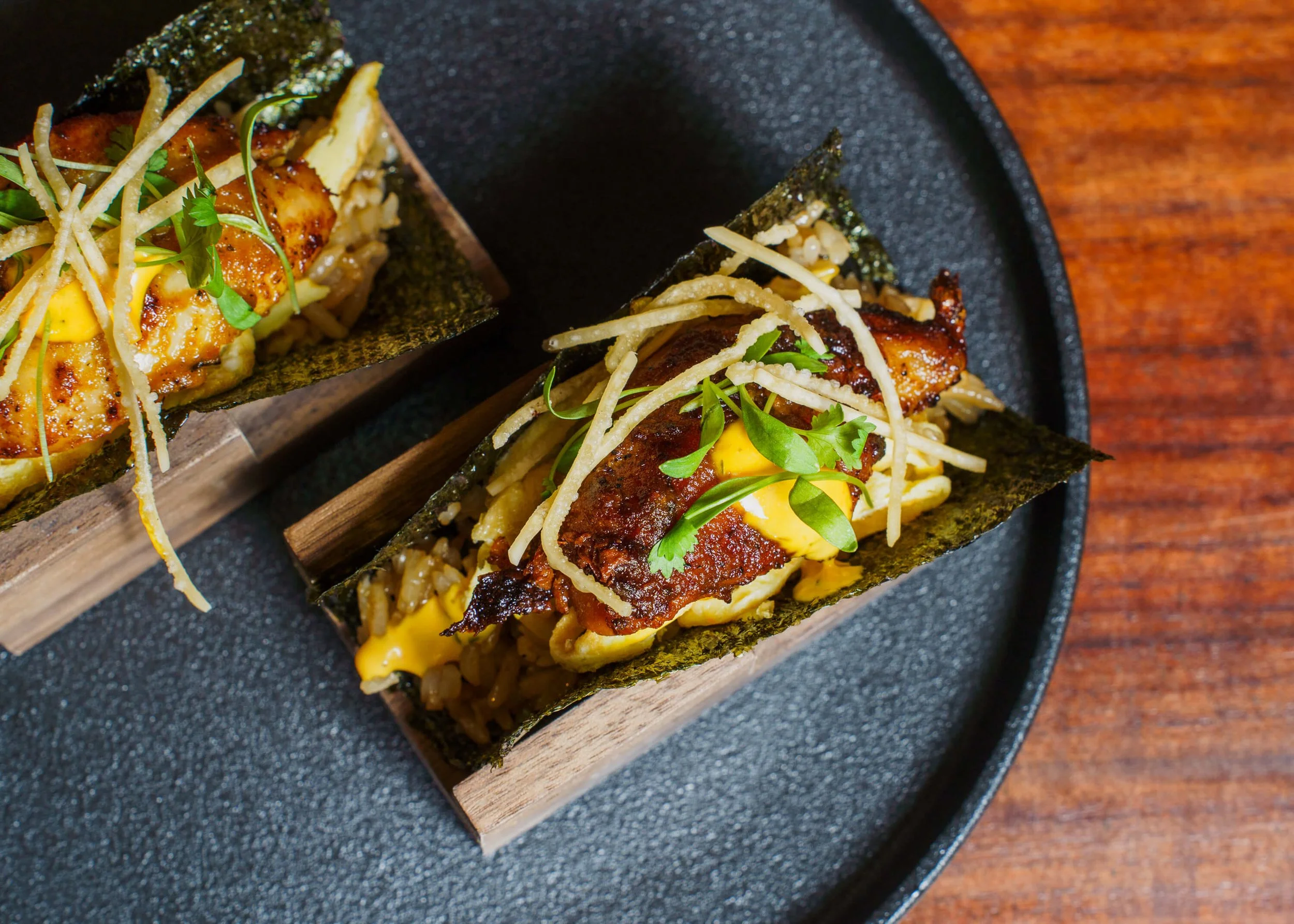 Close-up of two sushi tacos with grilled fish, shredded cheese, green herbs, and nori seaweed wrapping, served on a dark plate on a wooden table.