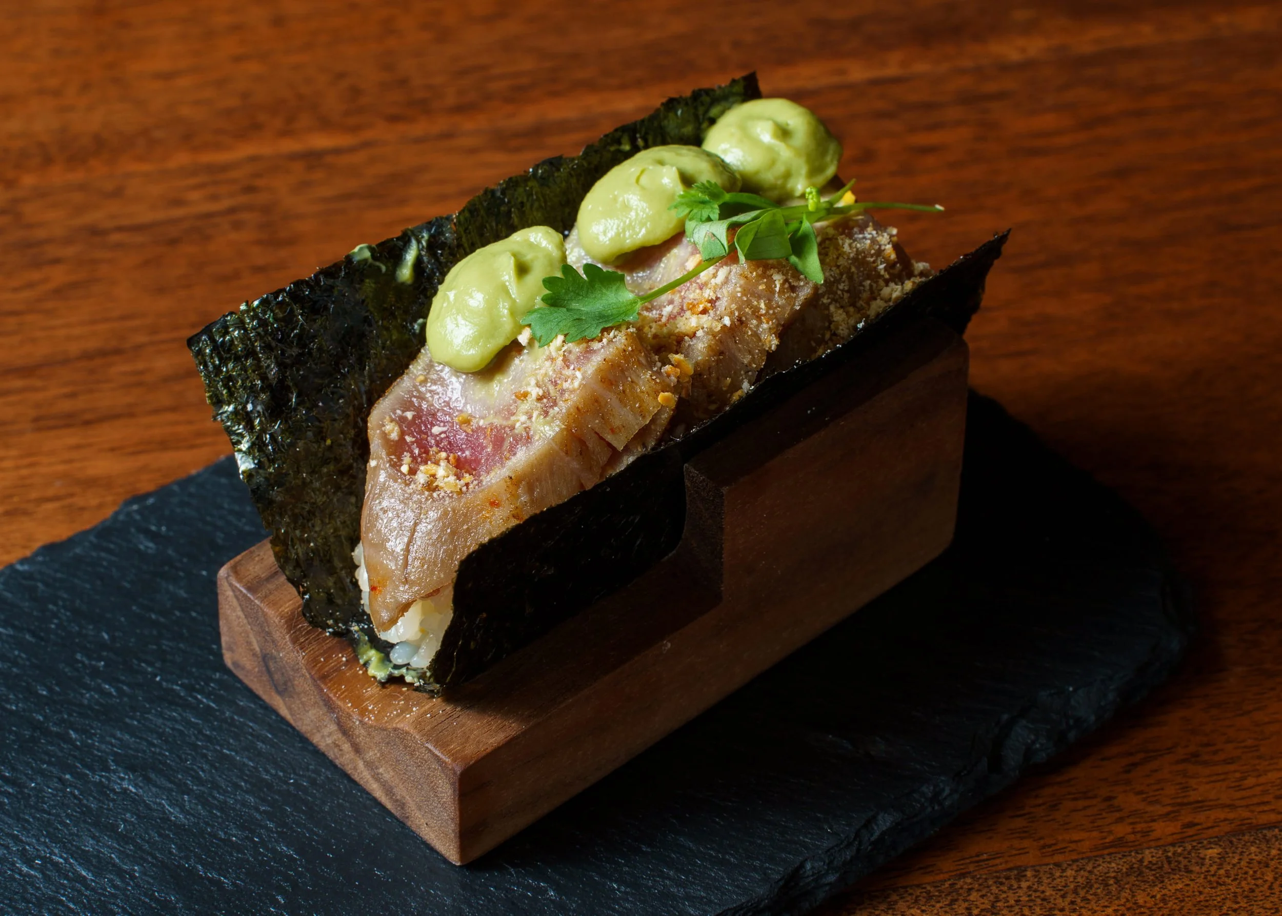 Close-up of a gourmet sushi roll topped with green sauce, garnished with green herbs, on a black slate plate on a wooden surface.