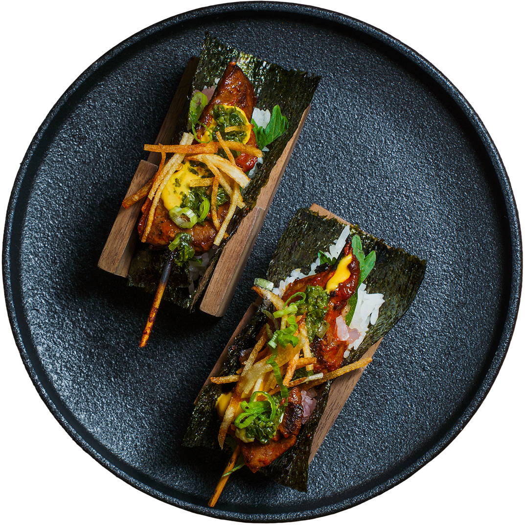 Two sushi handrolls wrapped in seaweed and placed on a black plate, topped with green onions, crispy potatoes, and sauce.