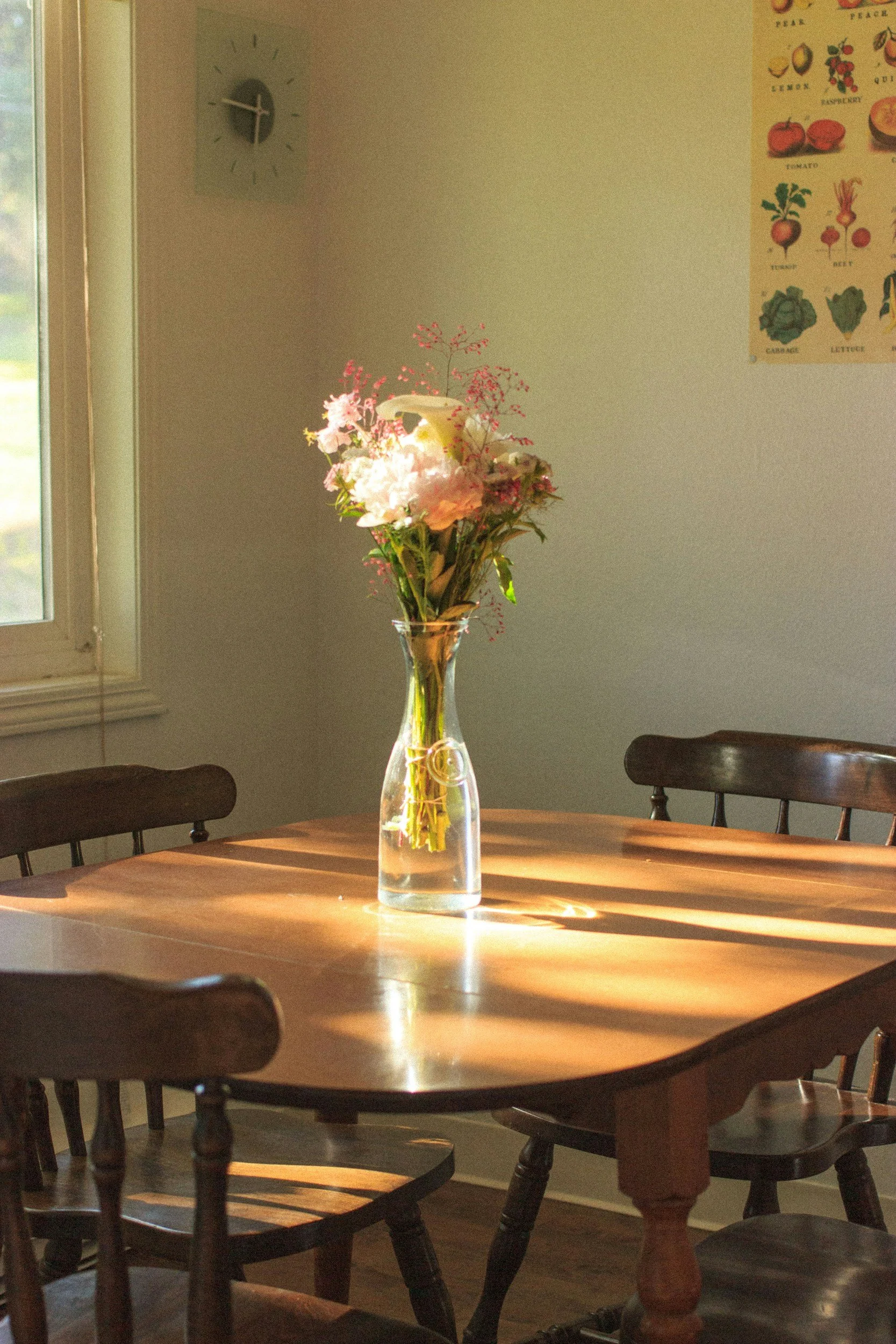 A round wooden dining table with wooden chairs around it and a tall glass vase with pink and white flowers on top. Sunlight is casting shadows on the table. In the background, a wall with a clock, a fruit chart, and a window.