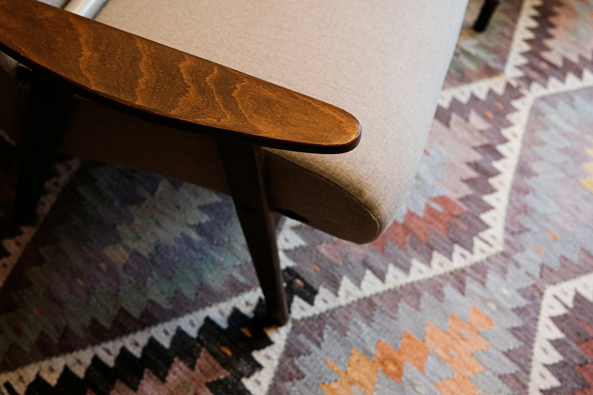 Close-up of a wooden chair armrest next to a beige upholstered chair and a patterned rug with zigzag and diamond shapes in various colors.