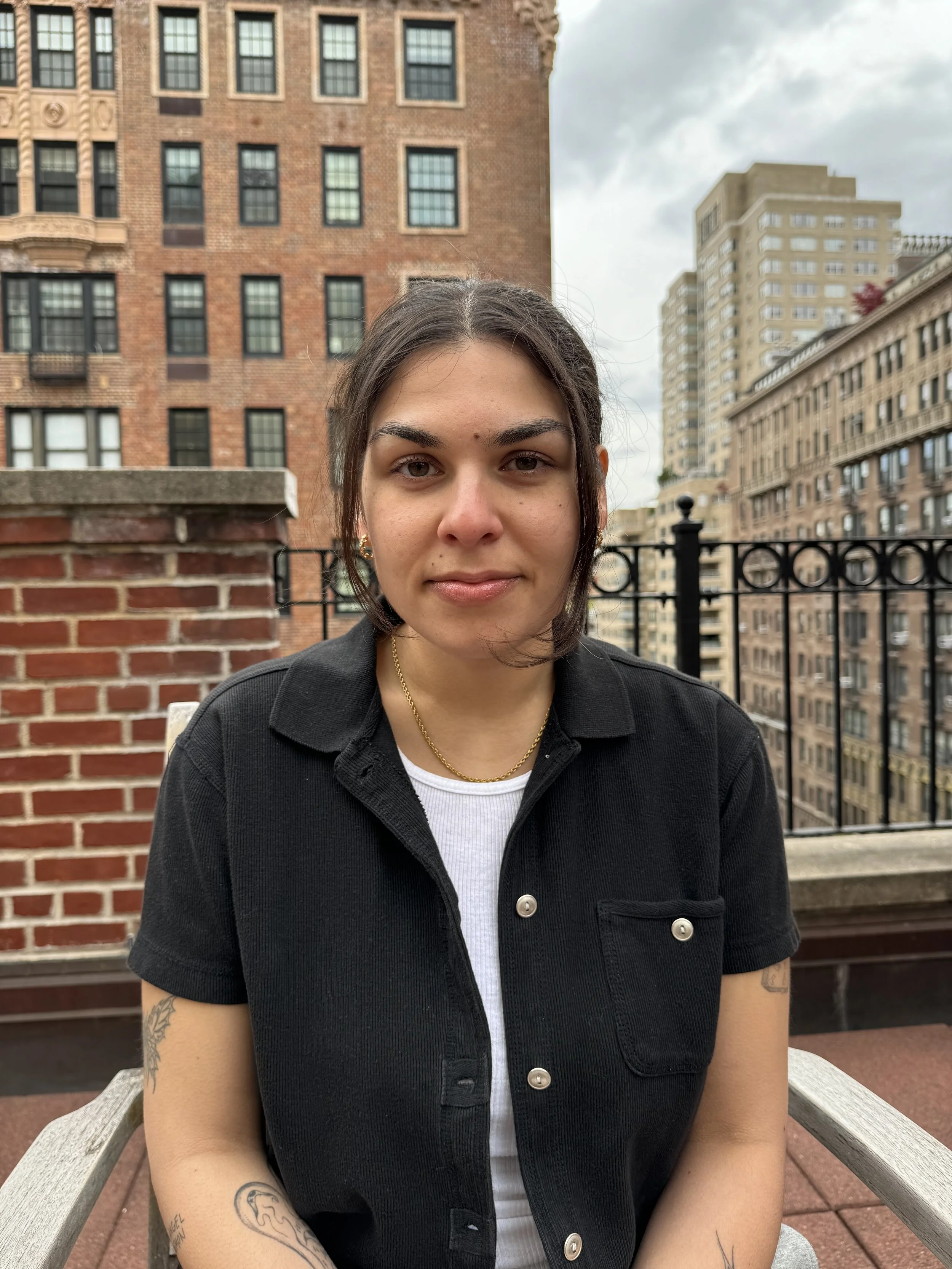 A young woman with short dark hair, wearing a black denim jacket, gold necklace, and earrings, sitting on an outdoor bench in an urban area with brick and concrete buildings in the background.
