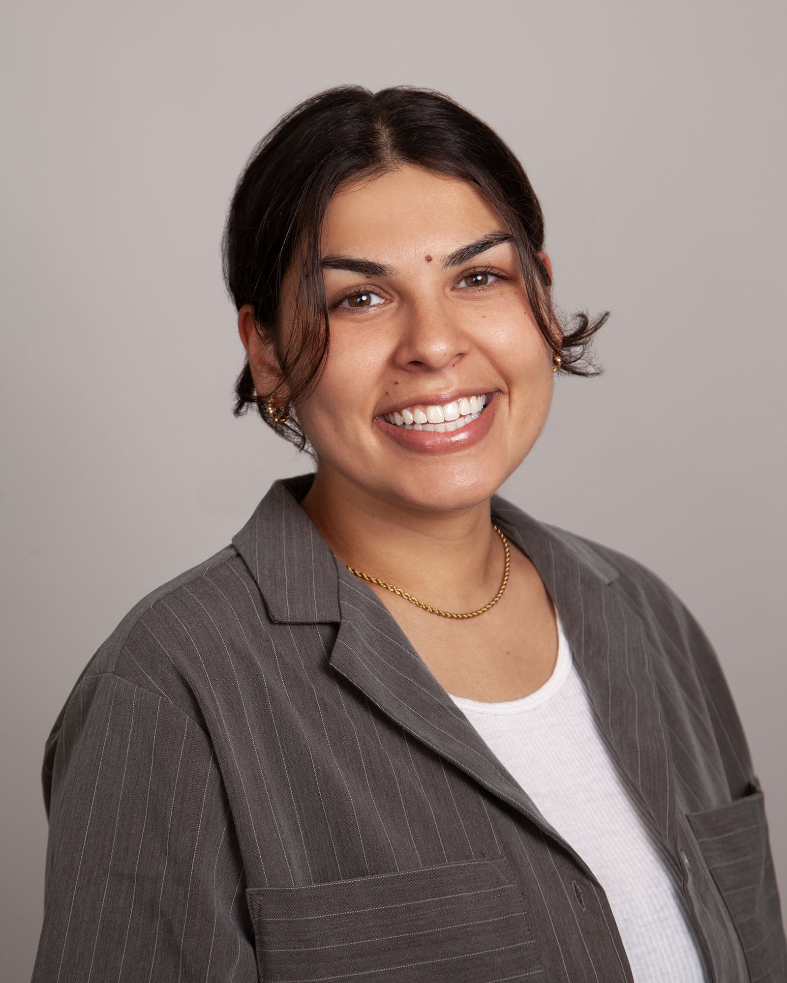 A woman with brown hair, wearing a gray pinstripe blazer, white shirt, gold necklace, and earrings, smiling at the camera against a neutral gray background.