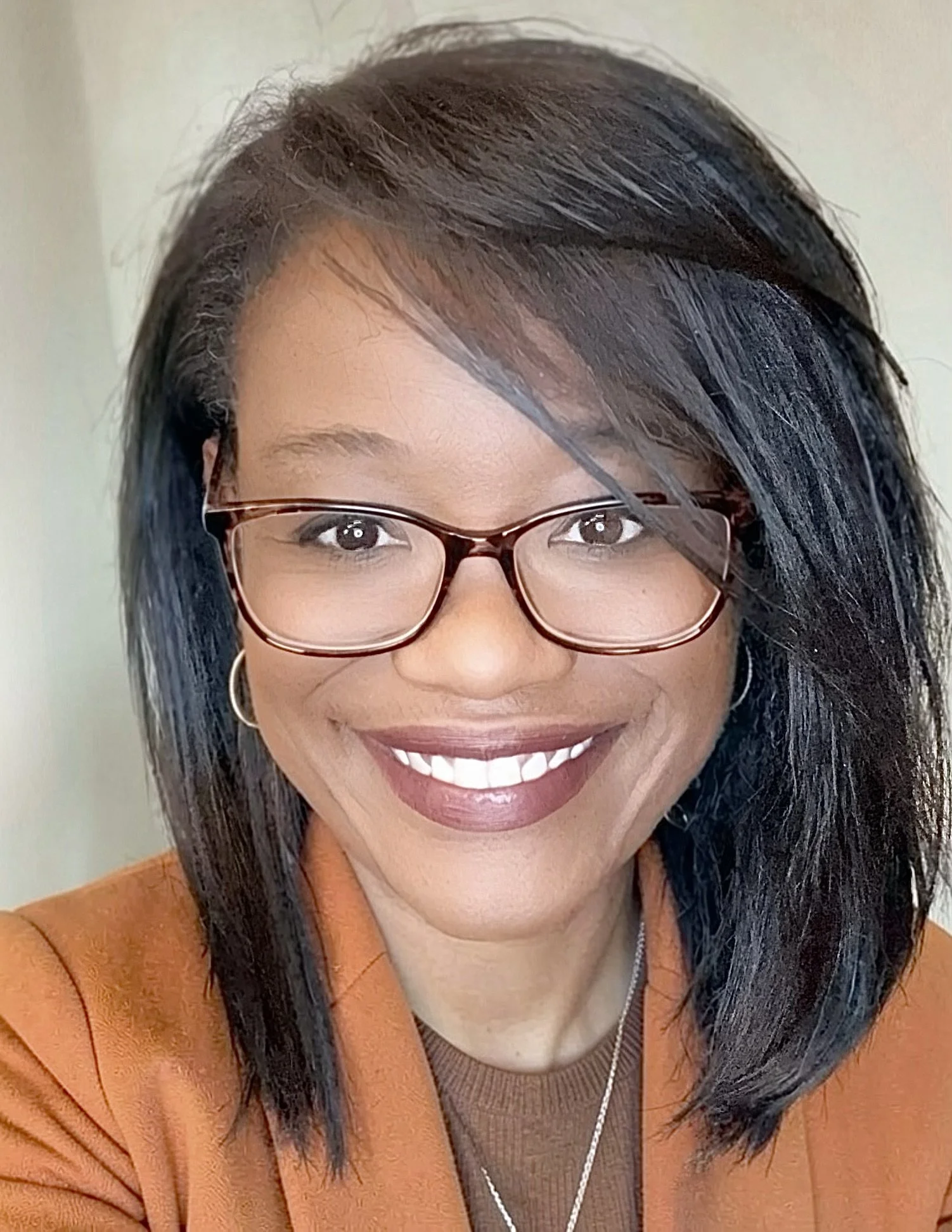A woman with dark, straight hair and glasses, smiling, wearing a brown jacket.