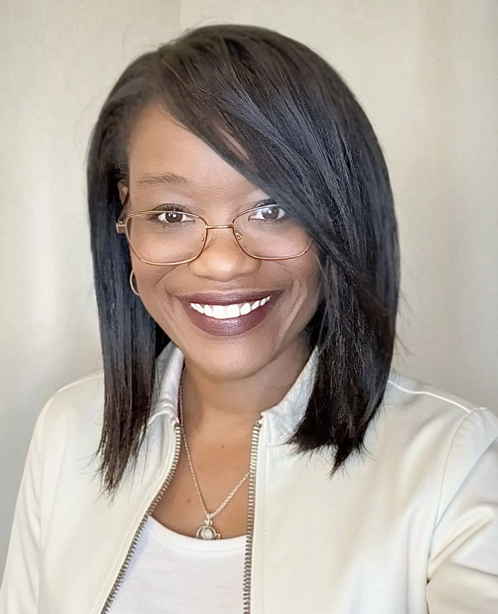 Close-up of a woman with dark, straight hair, wearing glasses, a white jacket, and jewelry, smiling against a plain background.