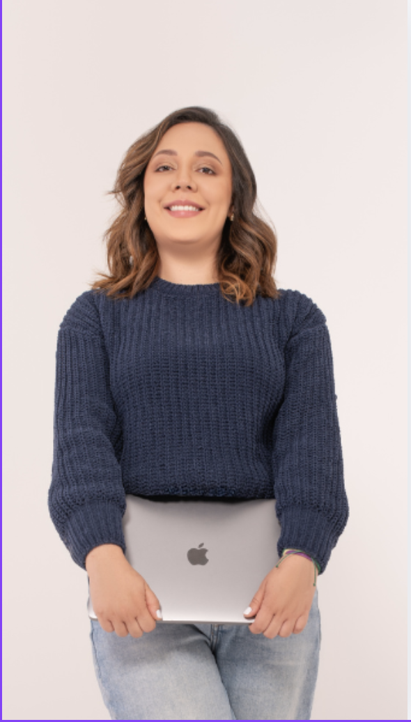 A woman with shoulder-length wavy hair, wearing a navy blue sweater and light jeans, smiling and holding a silver Apple laptop in front of her.