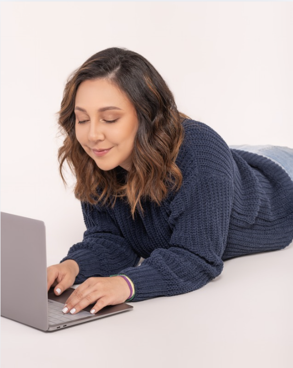 Young woman with wavy brown hair lying on her stomach and using a laptop.