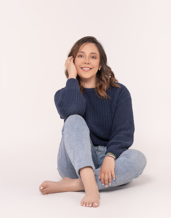 A young woman sitting cross-legged on the floor, smiling, with her left hand touching her hair. She has shoulder-length brown hair, wears a navy blue sweater and light blue jeans, and has a colorful bracelet on her right wrist. The background is plain white.