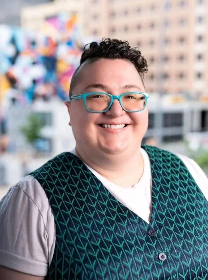 Person with short dark hair, wearing teal glasses, a white T-shirt, and a patterned vest, smiling in front of a blurred cityscape background with a colorful mural.