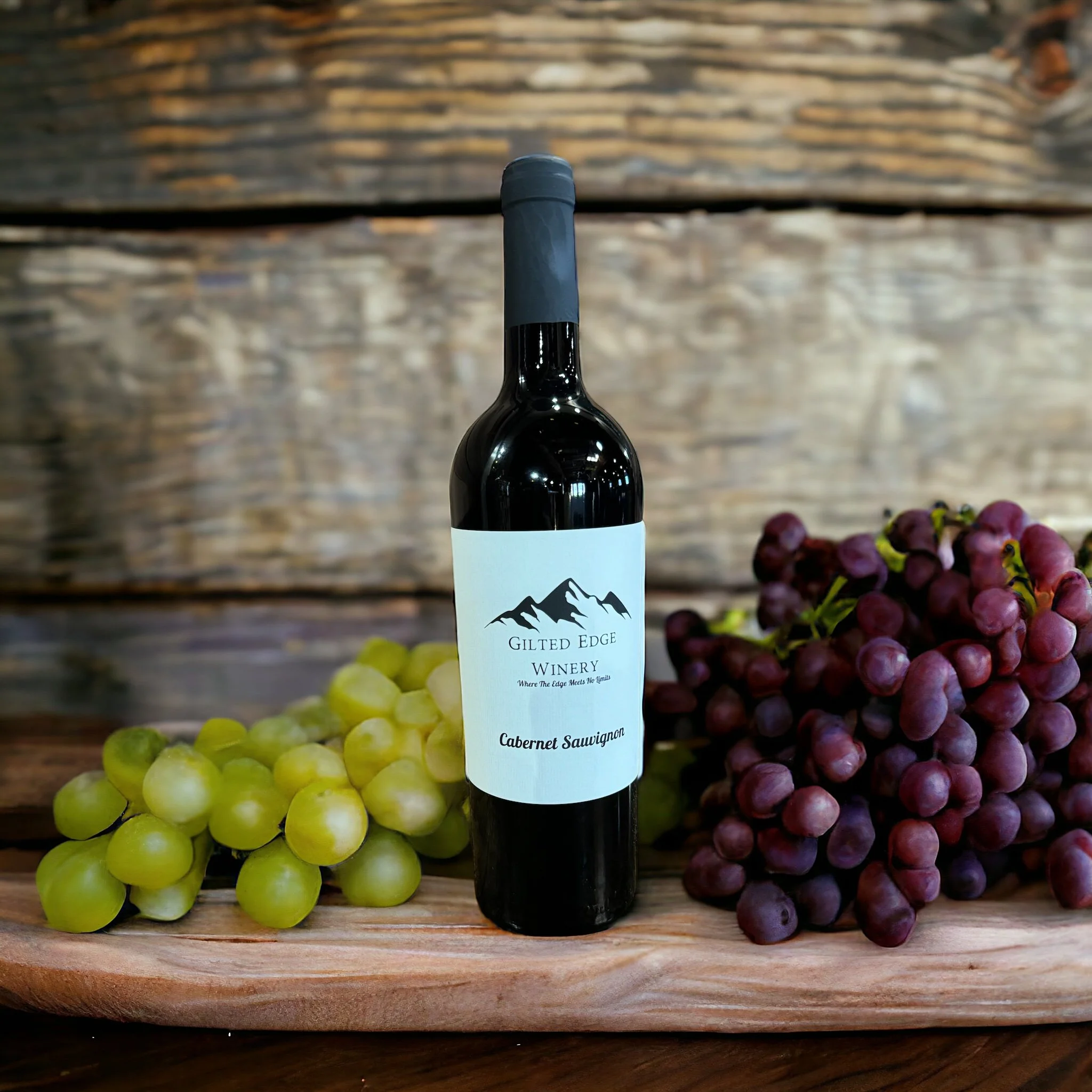 A bottle of wine with a mountain logo and the words 'Gilded Edge Winery' on the label, surrounded by green and purple grapes on a wooden surface.