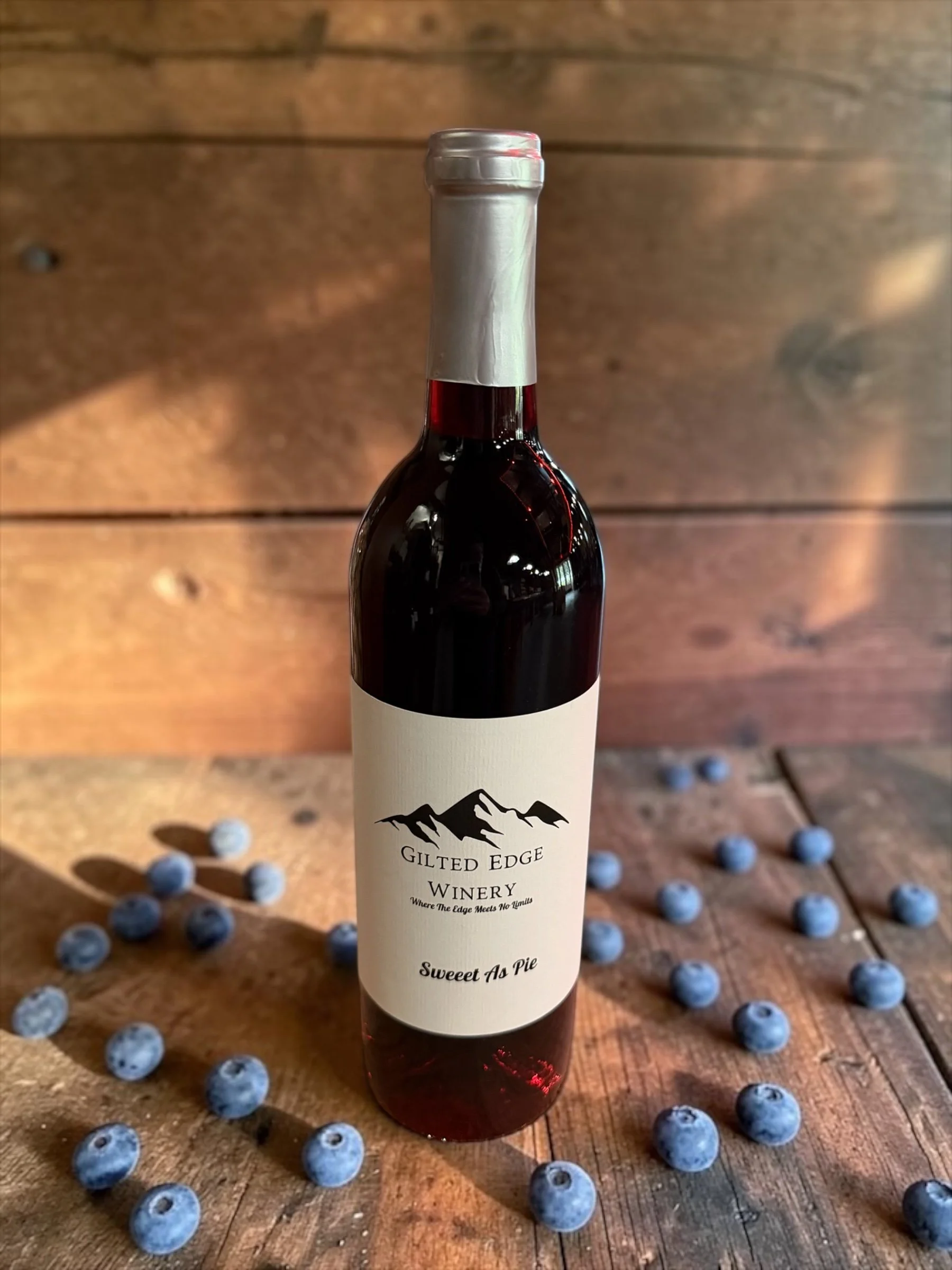 A bottle of wine labeled 'Gilted Edge Winery' with a mountain logo and the phrase 'Sweet As Pie' on a wooden surface. Blueberries are scattered around the bottle.