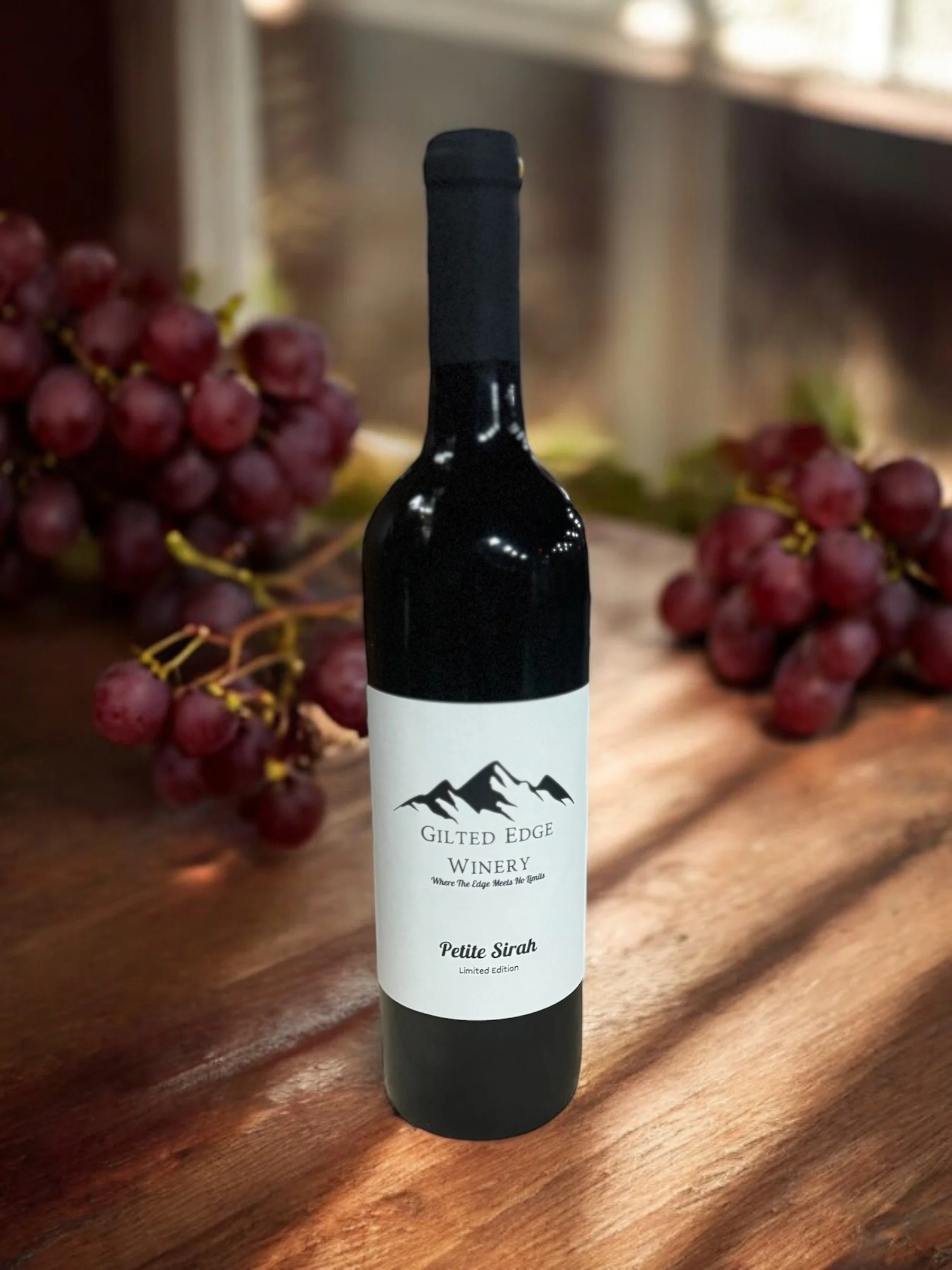A wine bottle with a white label featuring a mountain graphic, placed on a wooden surface with bunches of red grapes in the background.